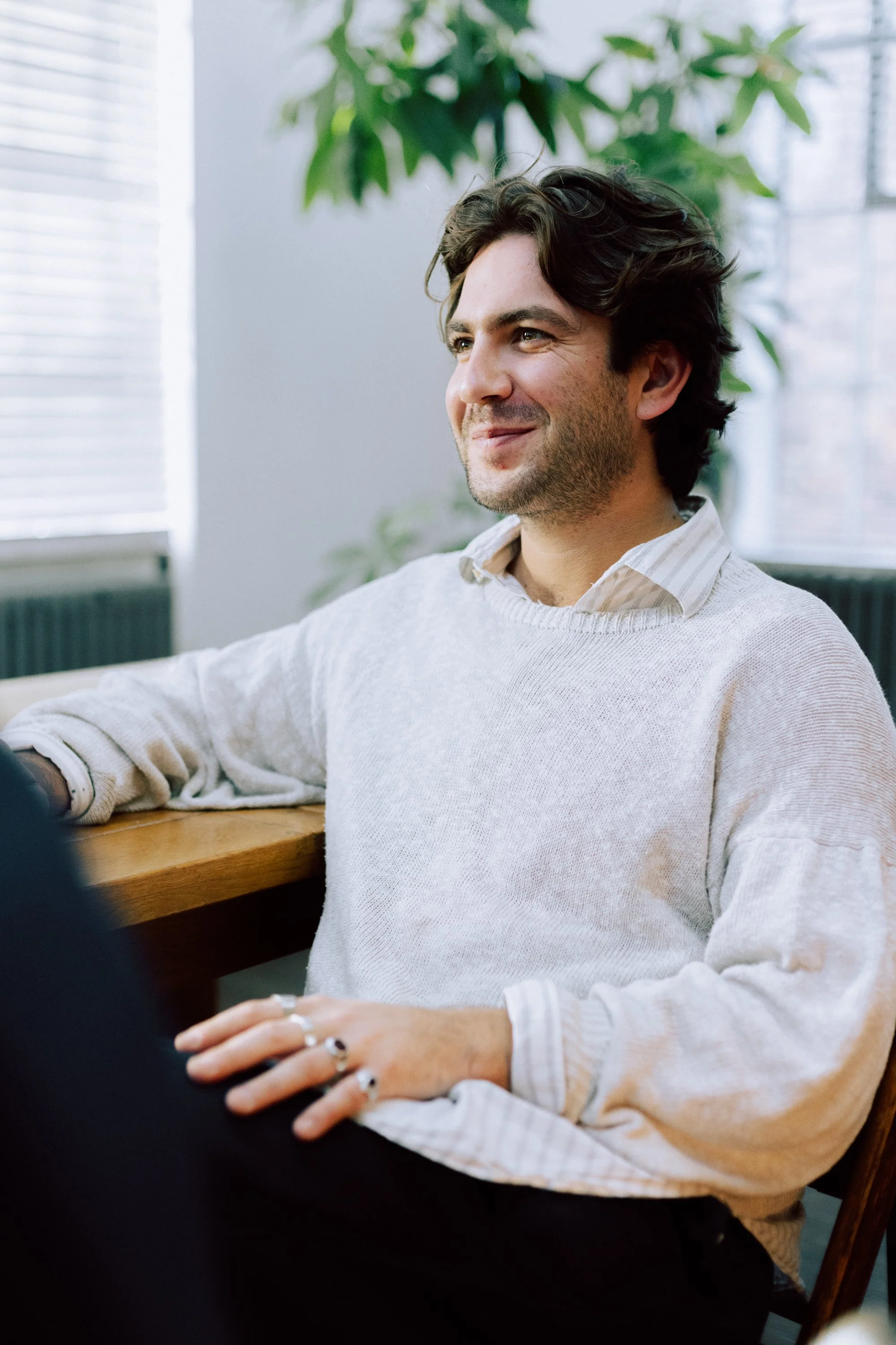 A man with dark hair, wearing a white sweater over a collared shirt, sitting at a wooden table, smiling, with green plants and window blinds in the background.