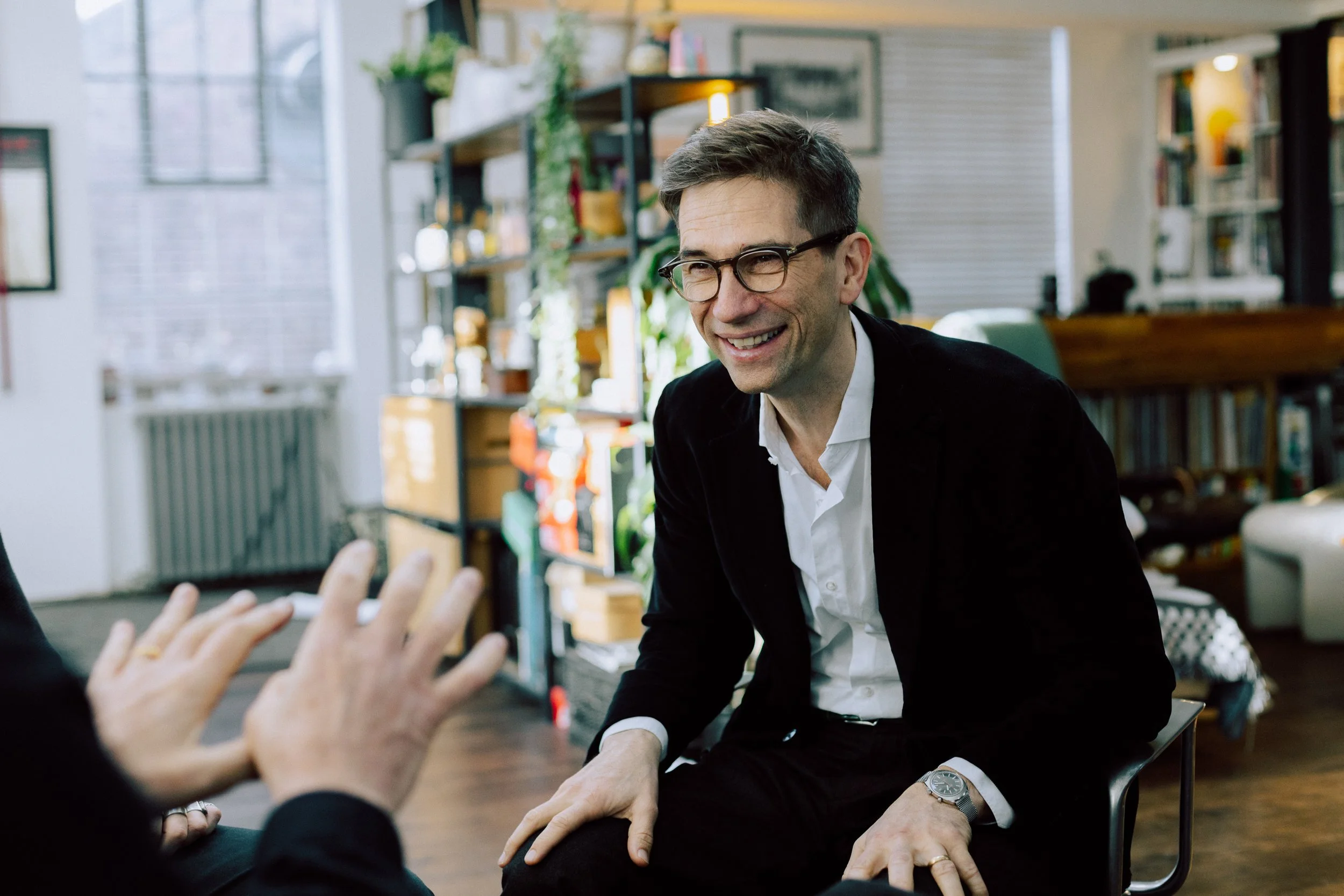 A man wearing glasses and a black blazer smiling during a conversation in a cozy, well-decorated room with bookshelves and green plants.