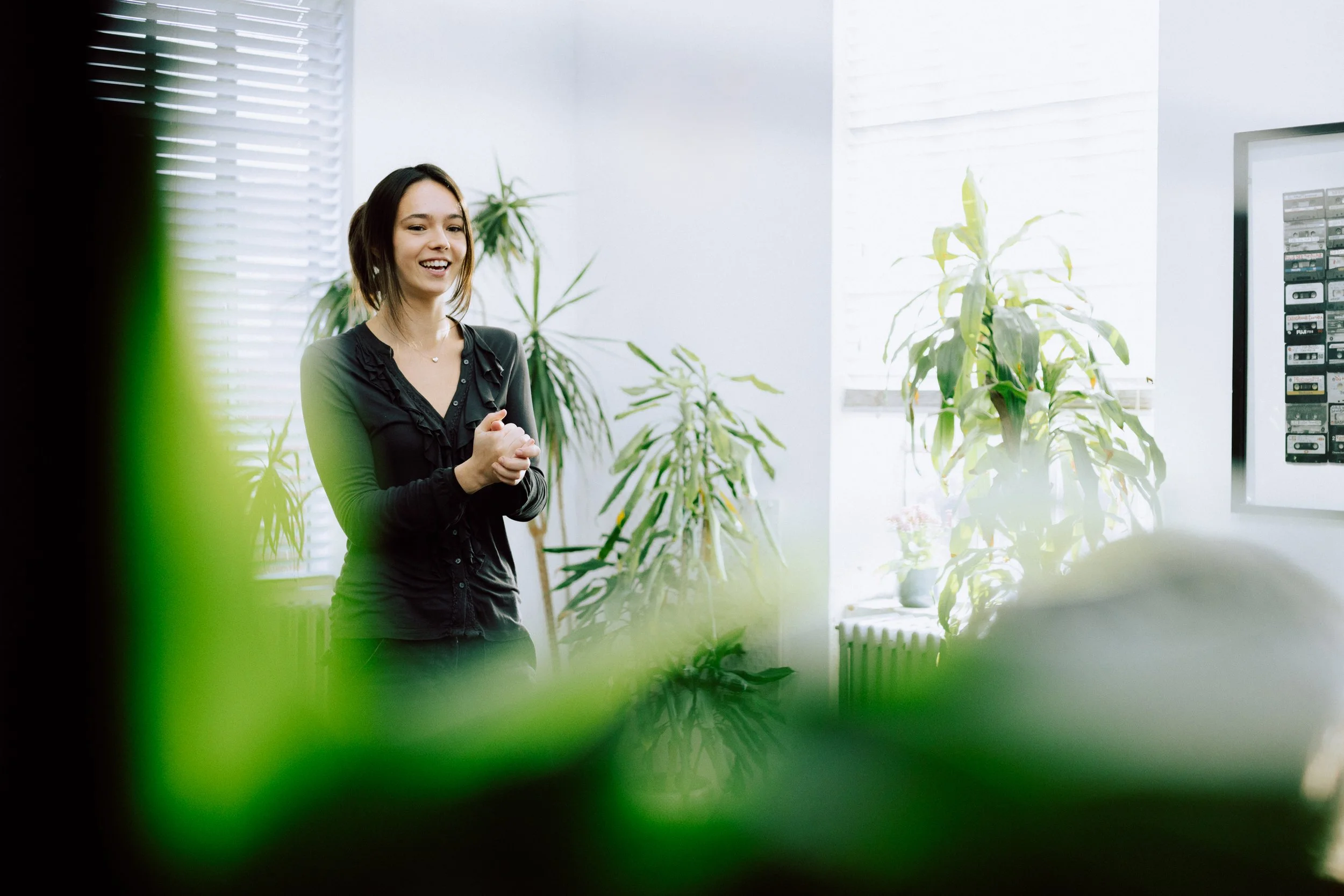 A young woman smiling and clapping her hands in a bright, modern room with large windows, green plants, and framed artwork on the wall.