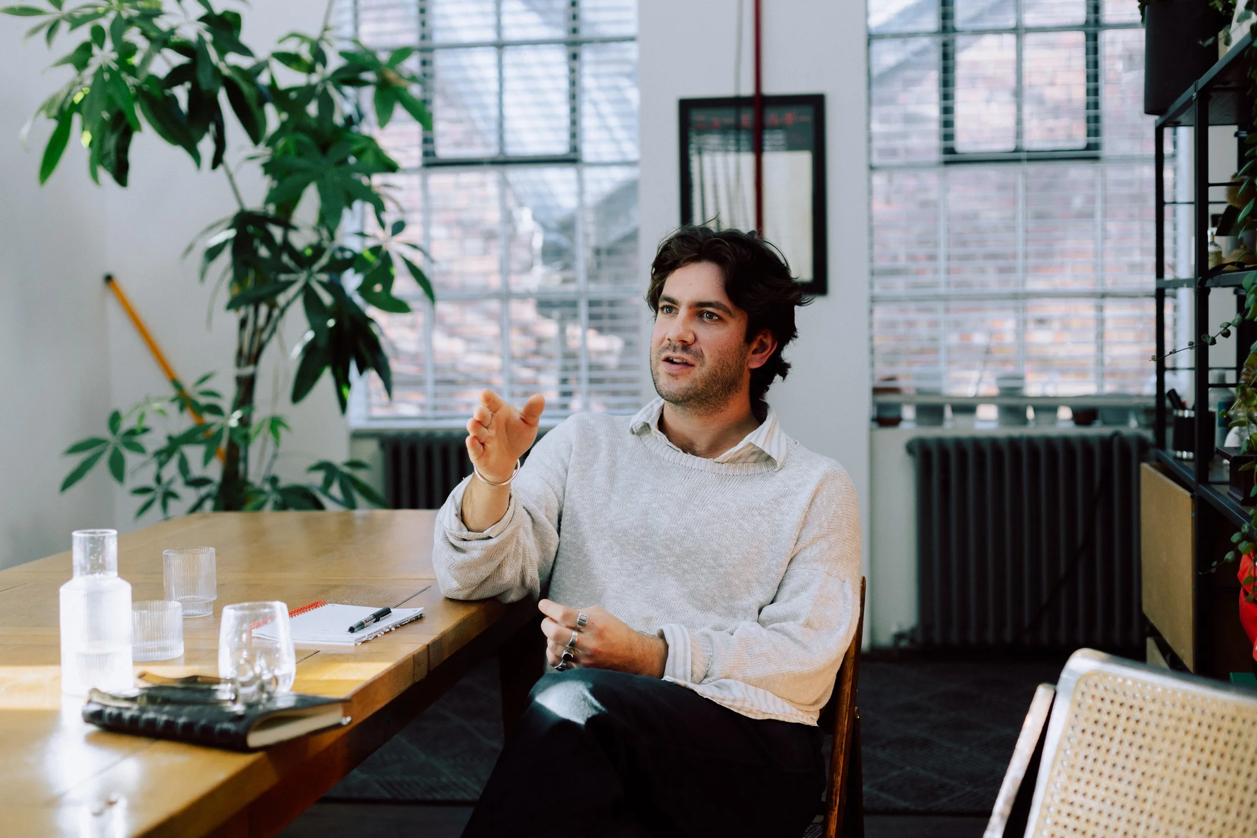 A man with dark wavy hair, wearing a light-colored sweater, sitting at a wooden table in a bright room with large windows and green plants, gesturing with his right hand.