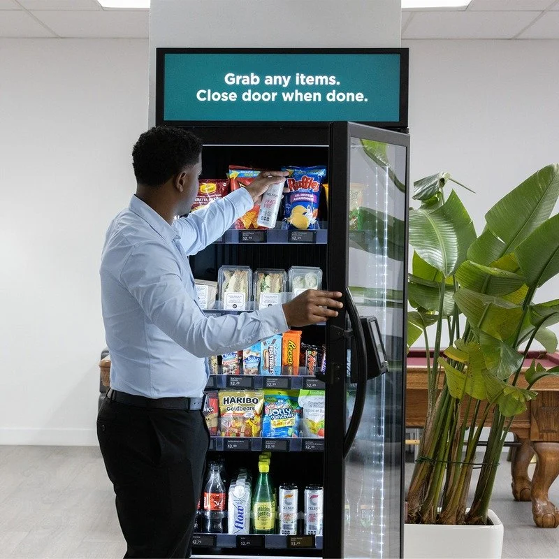 A man in a light blue shirt and black pants reaching into a vending machine filled with snacks and drinks, with a sign above that says, 'Grab any items. Close door when done.'