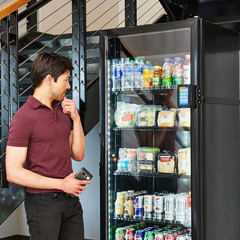 A young man with dark hair, dressed in a maroon polo and black pants, standing in front of a glass vending machine with snacks, drinks, and bottled water, in an indoor setting with a staircase and metal railing in the background.