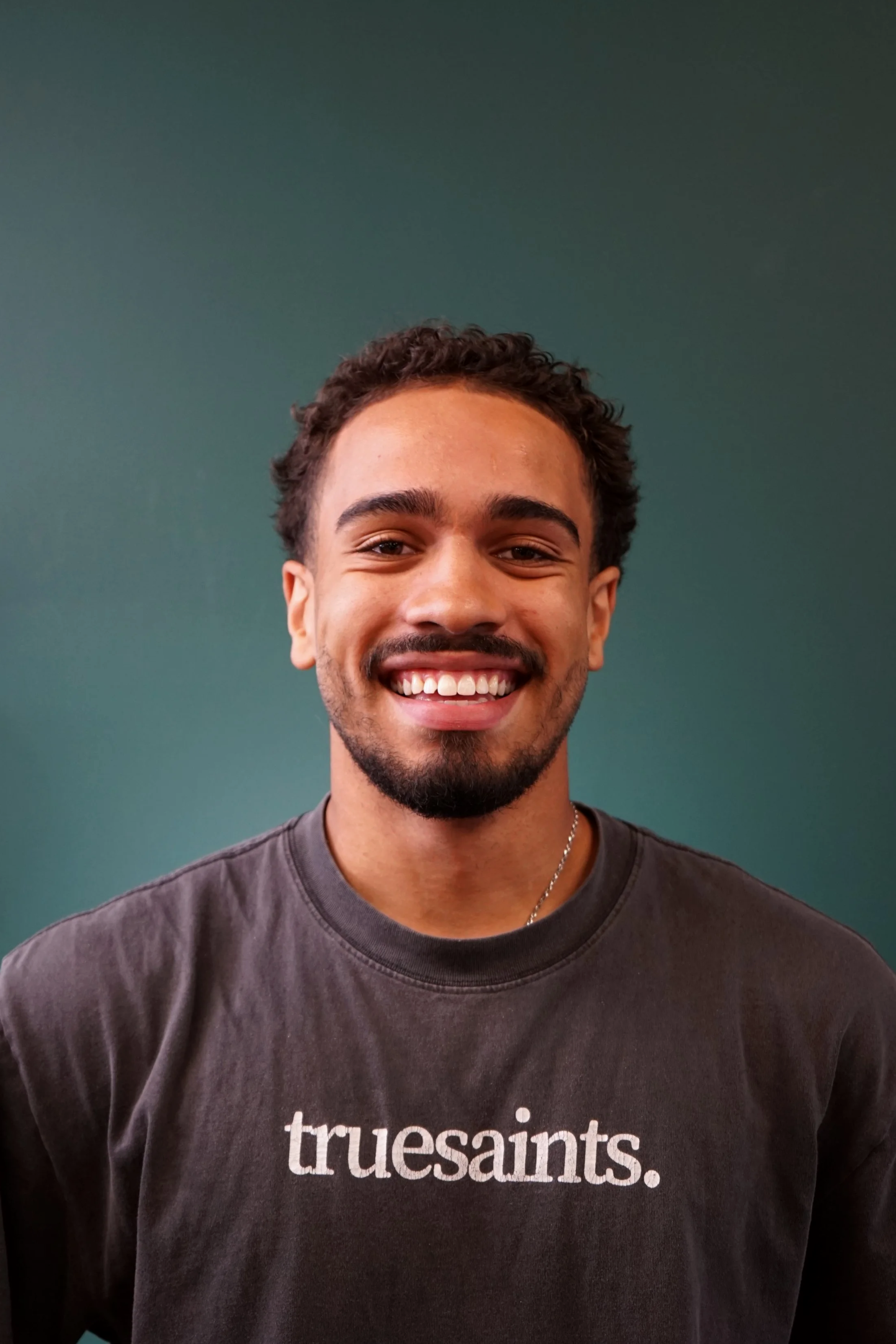 Adain Holliday, barber specializing in men's shear work at Shop 13 Barber Lounge in Bucks County, PA. Fairless Hills, PA. A Black and Spanish barber/hairstylist with short curly hair, a light beard, and a mustache smiling at the camera.