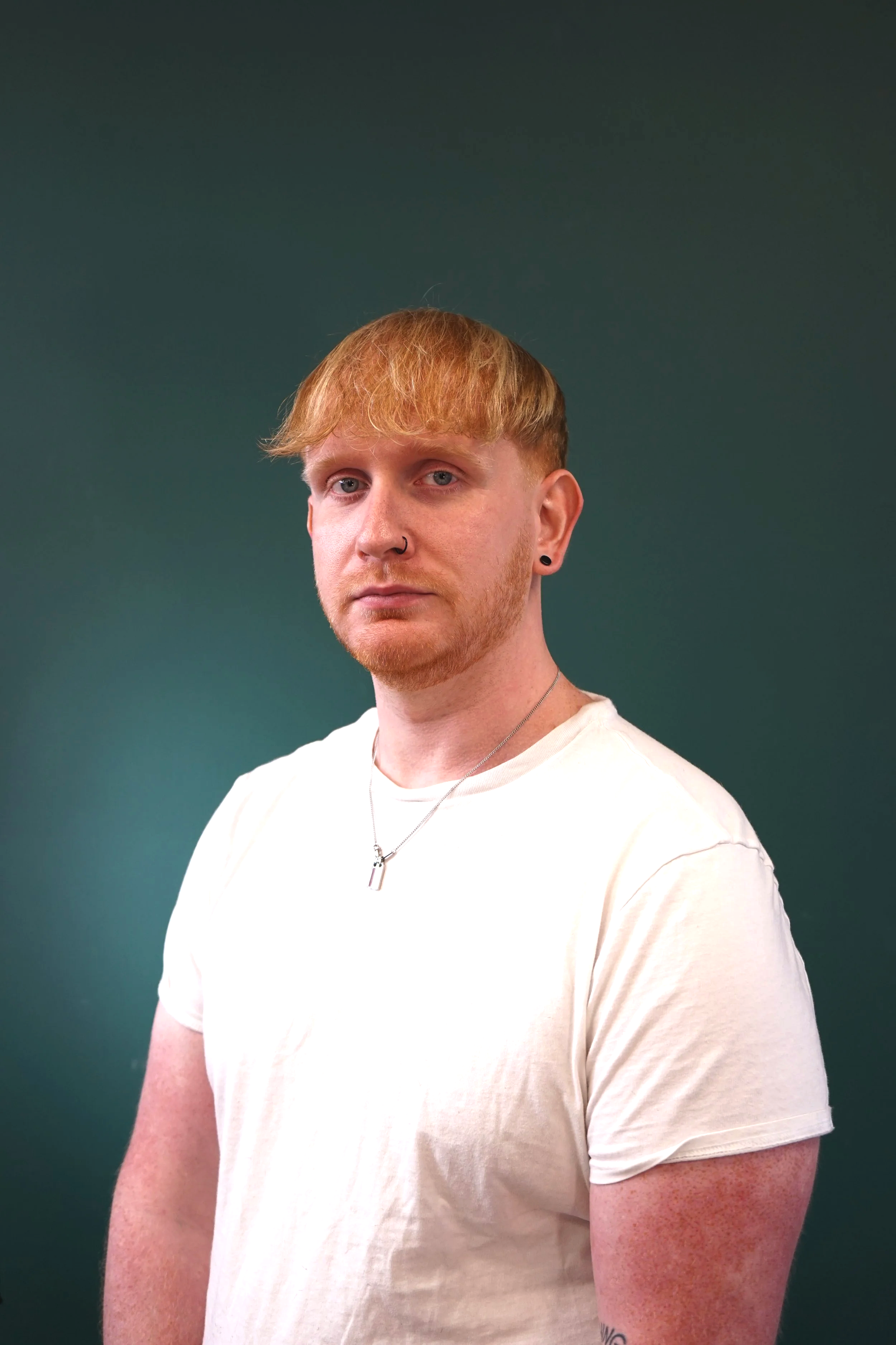 A well-connected barber with a beard and nose and ear piercings, wearing a white T-shirt and silver necklace, standing against a dark green background. Who likes, gives great fades, and does well with beards.