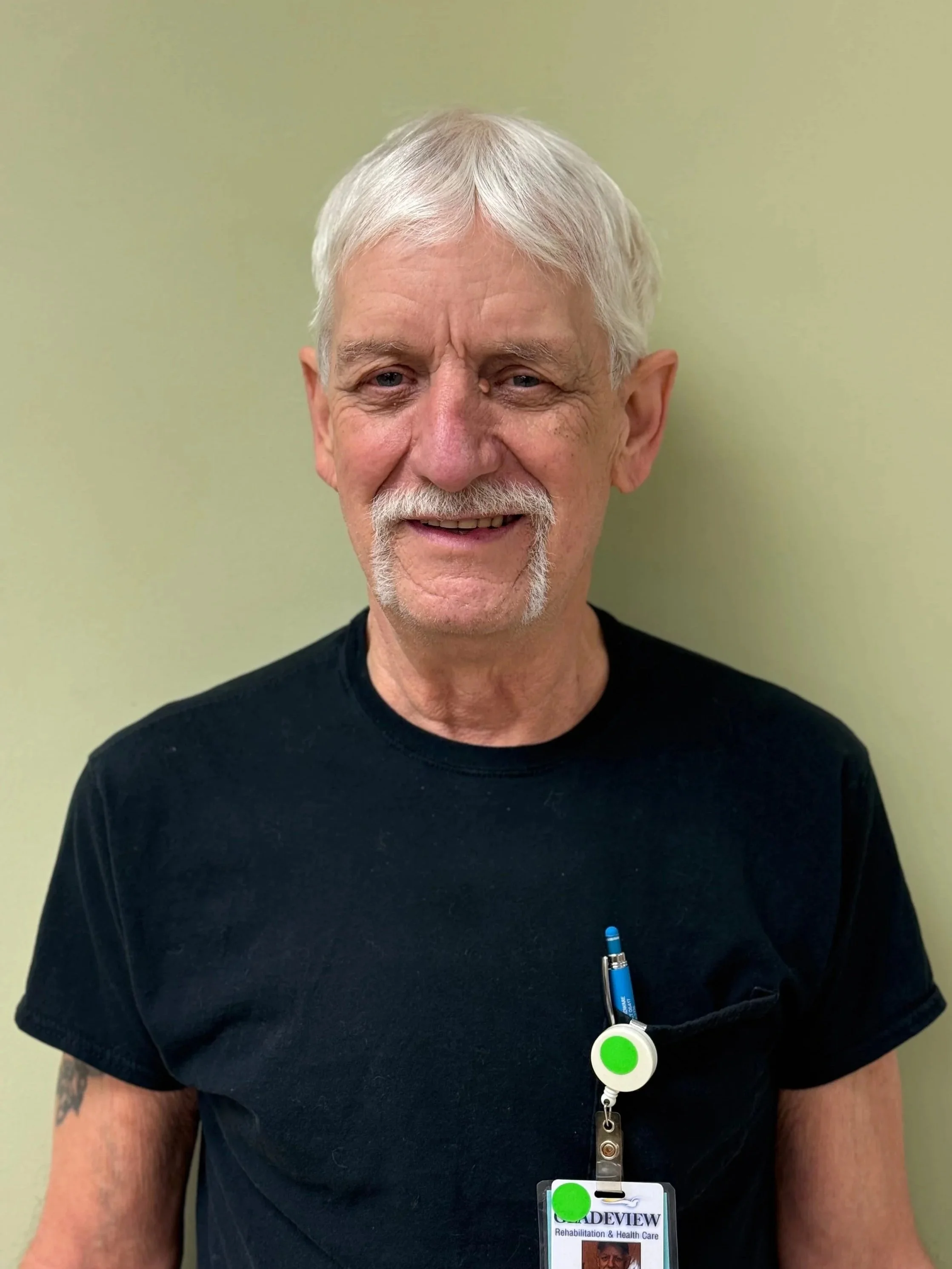 An older man with white hair, a mustache, and a goatee smiling, wearing a black t-shirt, with a badge and a pen in his shirt pocket, standing against a plain light green wall.