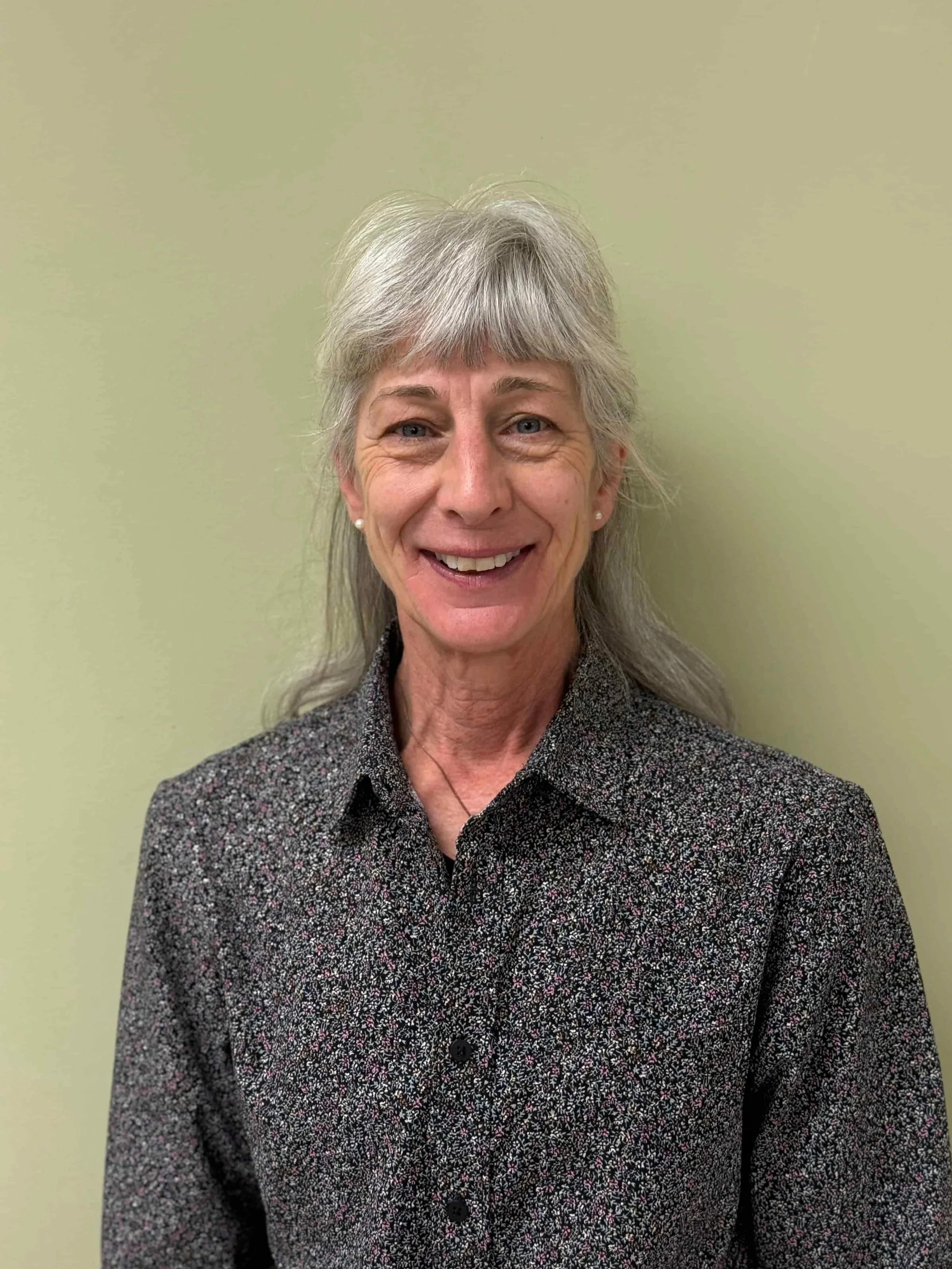 A smiling elderly woman with gray hair and pearl earrings, wearing a dark patterned shirt, standing against a light green wall.
