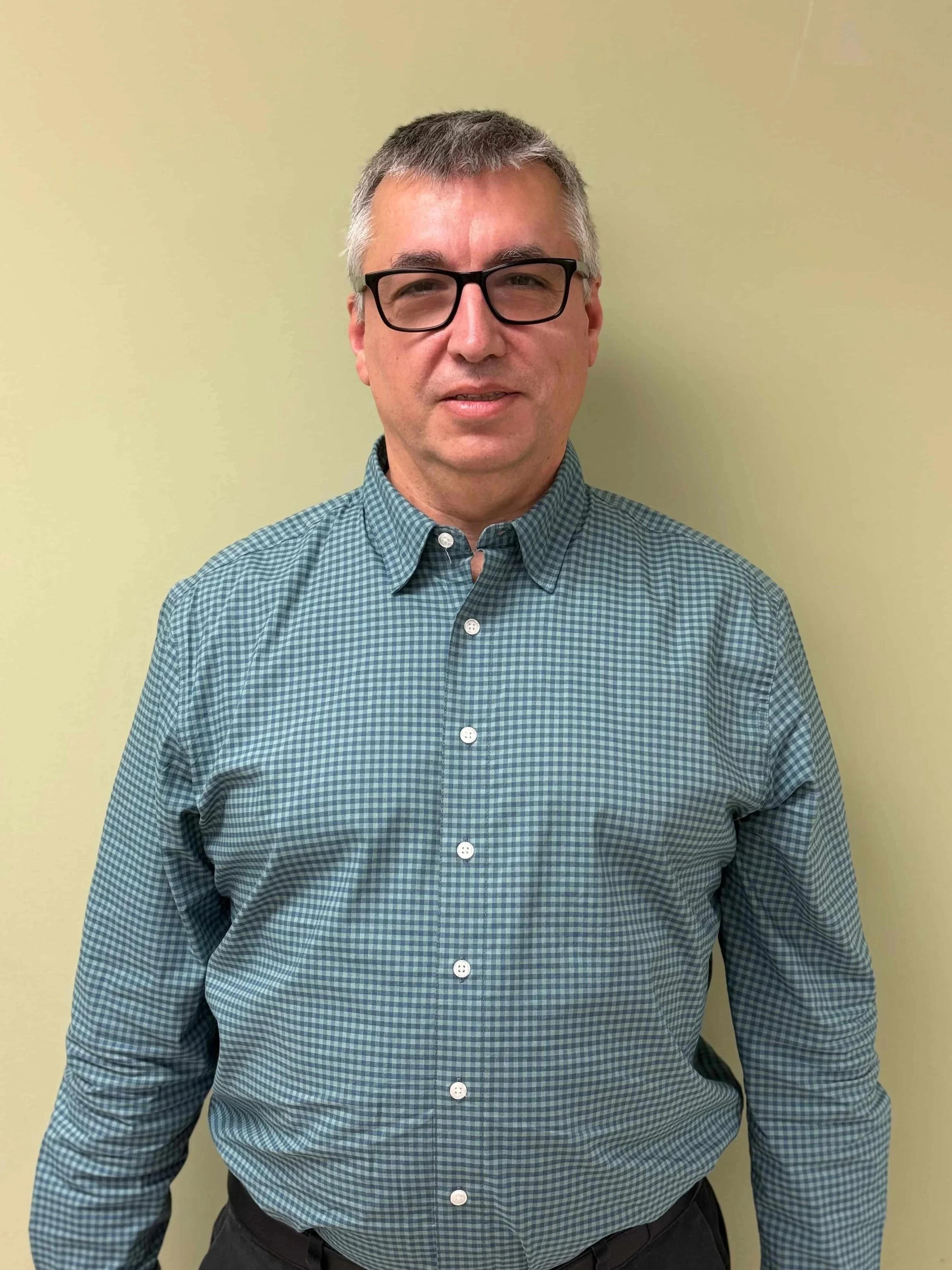 A middle-aged man with gray hair, wearing black glasses, and a green checkered shirt standing against a light green wall.