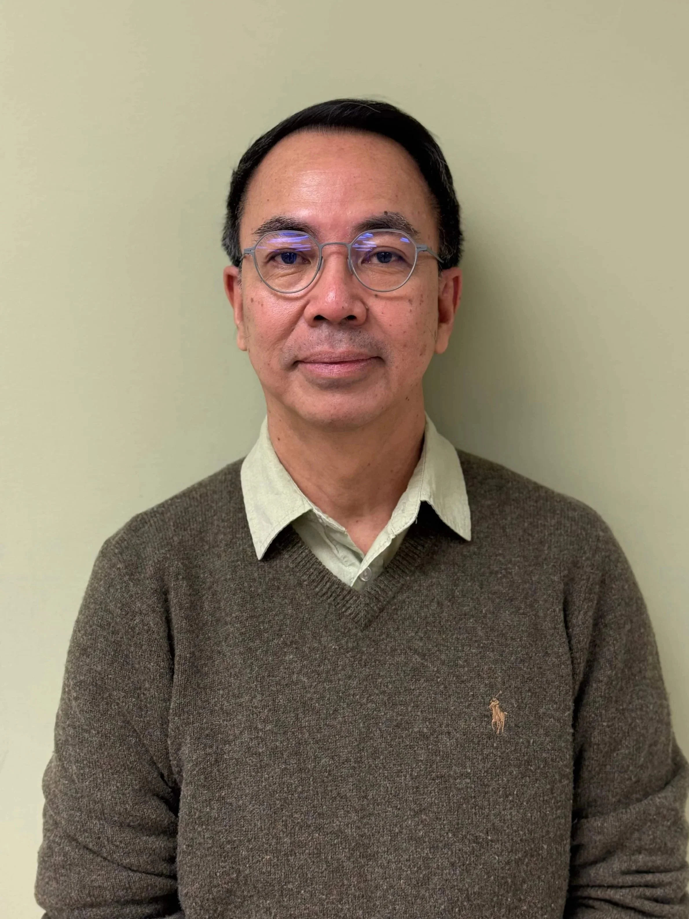 A middle-aged man with glasses, wearing a beige collared shirt underneath a brown sweater with a small embroidered logo, standing against a light green wall.