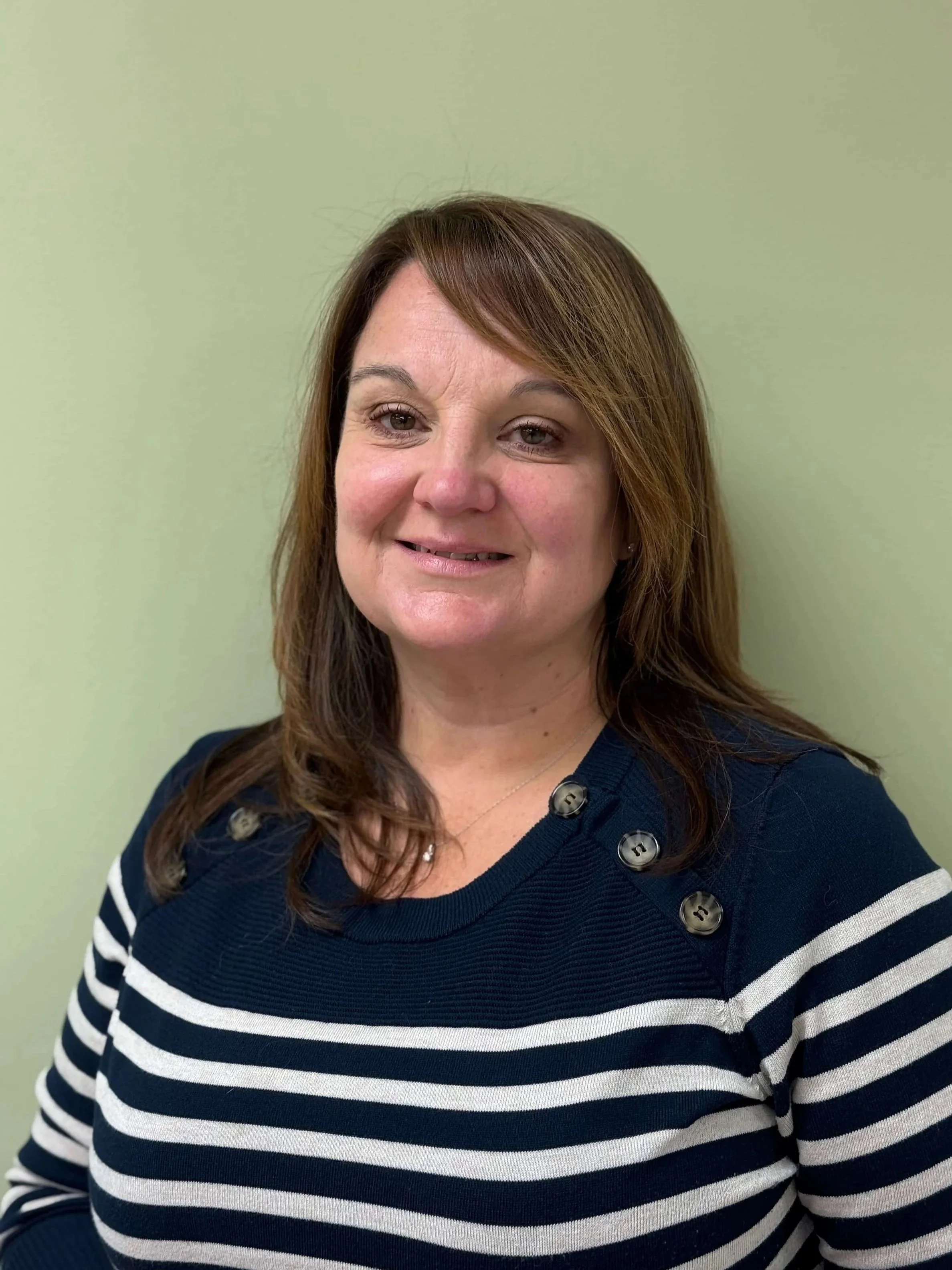 A woman with shoulder-length brown hair wearing a navy blue and white striped sweater with button details on the shoulder, standing against a light green wall.