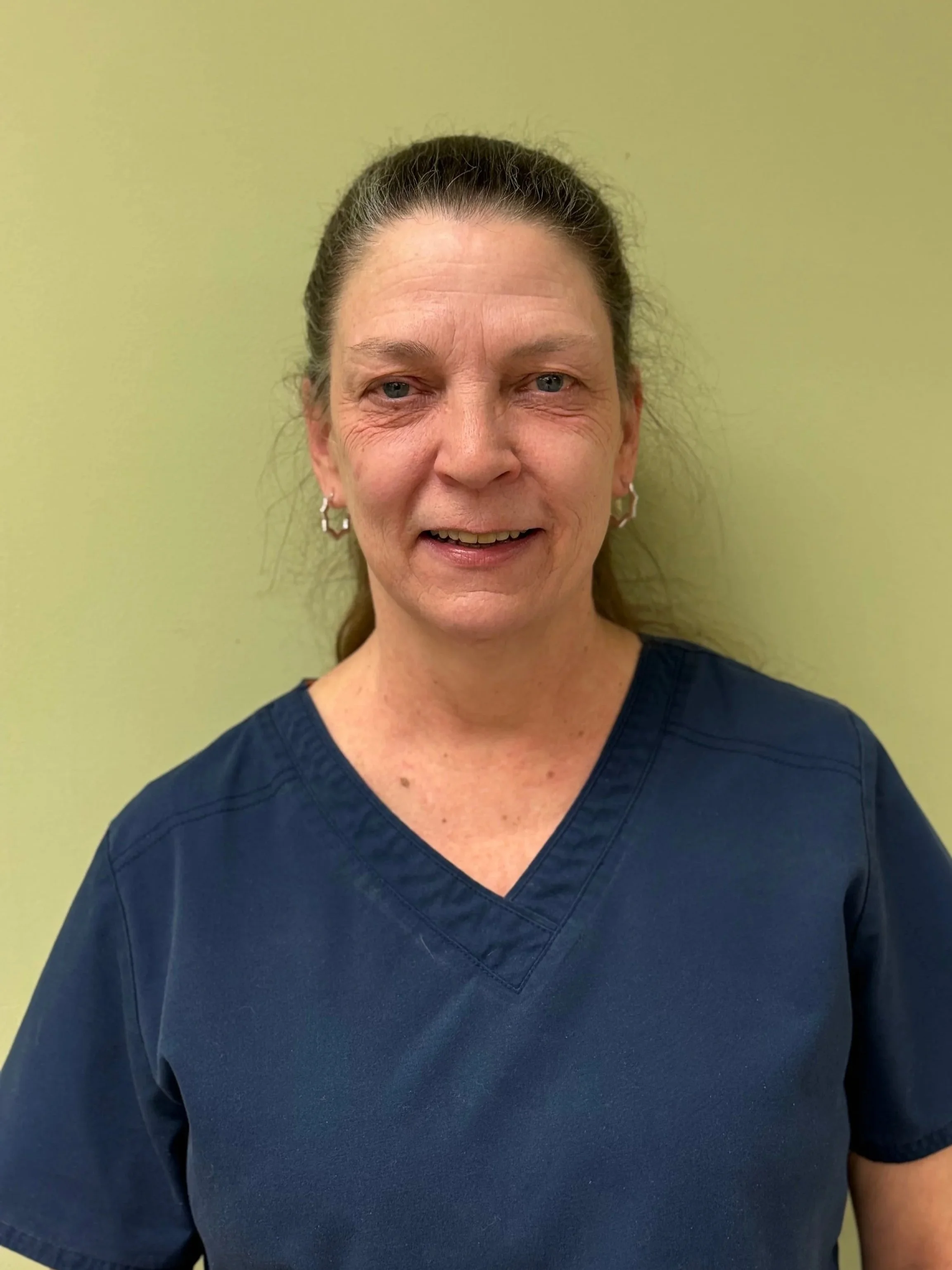 A middle-aged woman with brown hair tied back, wearing blue medical scrubs and star-shaped earrings, standing against a yellow-green wall.