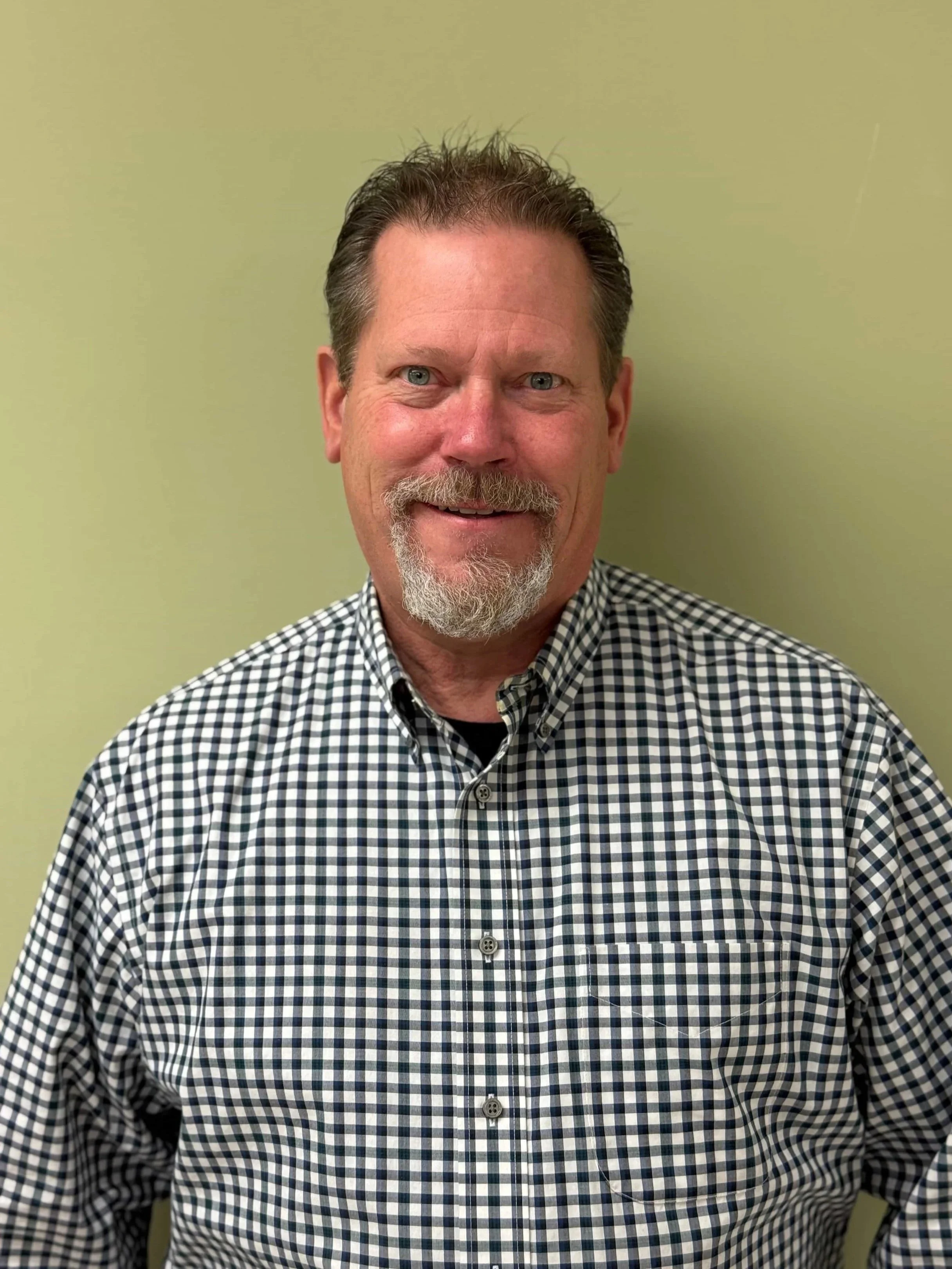 A middle-aged man with gray hair and a beard, wearing a checkered shirt, standing against a plain light green wall.