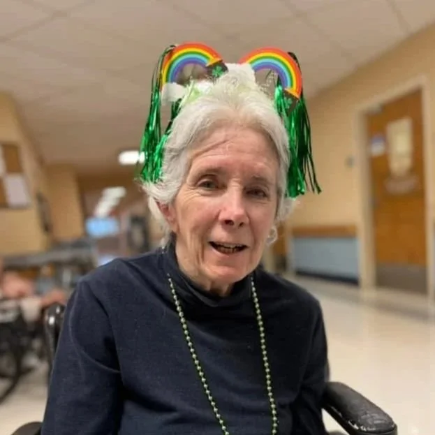 Older woman with white hair wearing a headband decorated with rainbows and green streamers, smiling and sitting in a wheelchair.