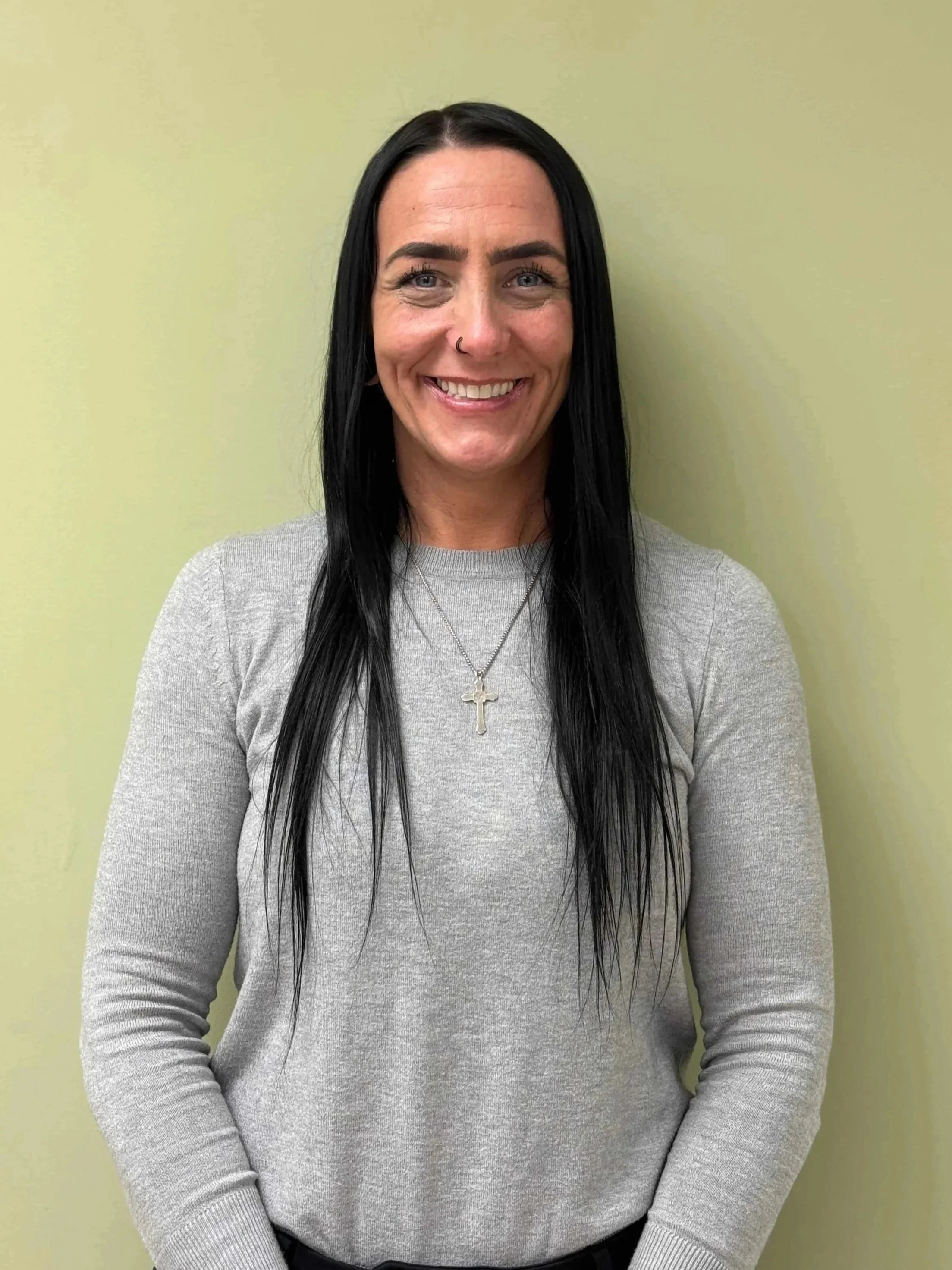 A woman with long black hair, wearing a light gray sweater, standing against a light green wall, smiling, with cross necklace and nose ring.
