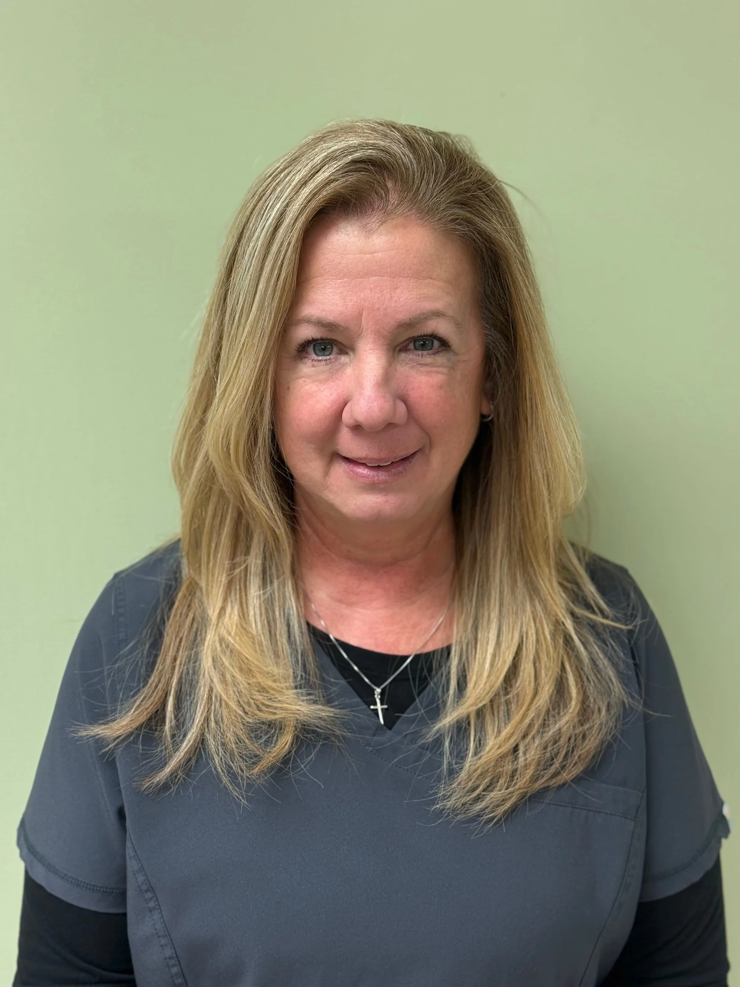 A woman with shoulder-length blonde hair wearing scrubs and a cross necklace, standing against a light green wall.