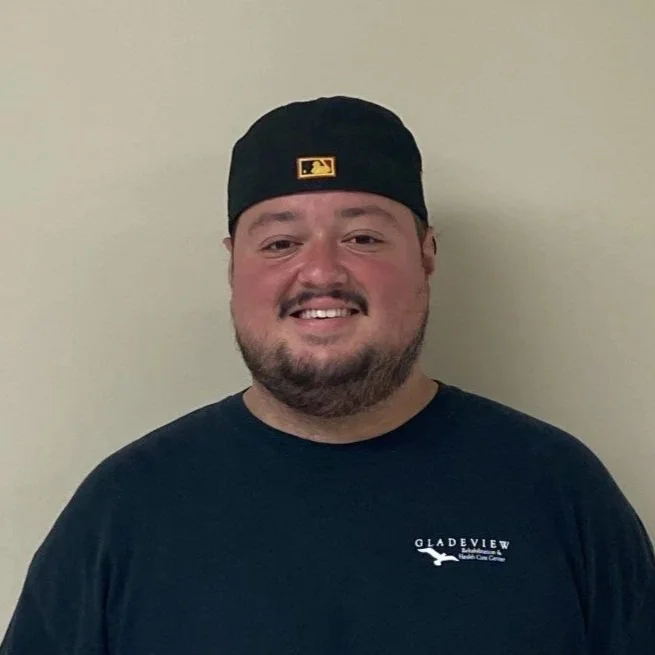 Smiling man wearing a black baseball cap with a yellow logo and a dark t-shirt with 'Gladeview' printed on it, standing against a plain light-colored wall.