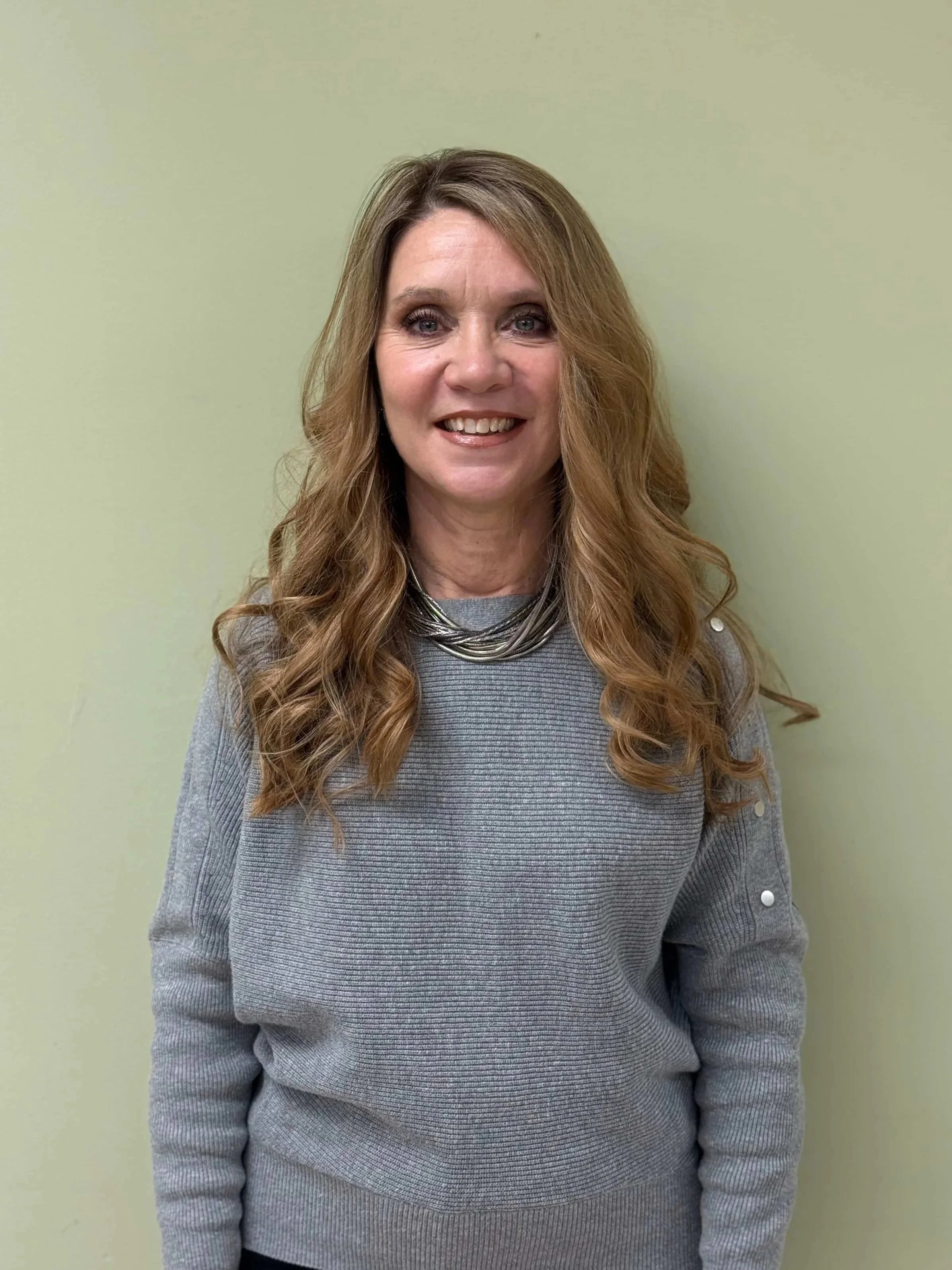 A woman with long, wavy, light brown hair, smiling, wearing a gray sweater with metallic buttons on the sleeves and a layered silver necklace, standing against a light green wall.