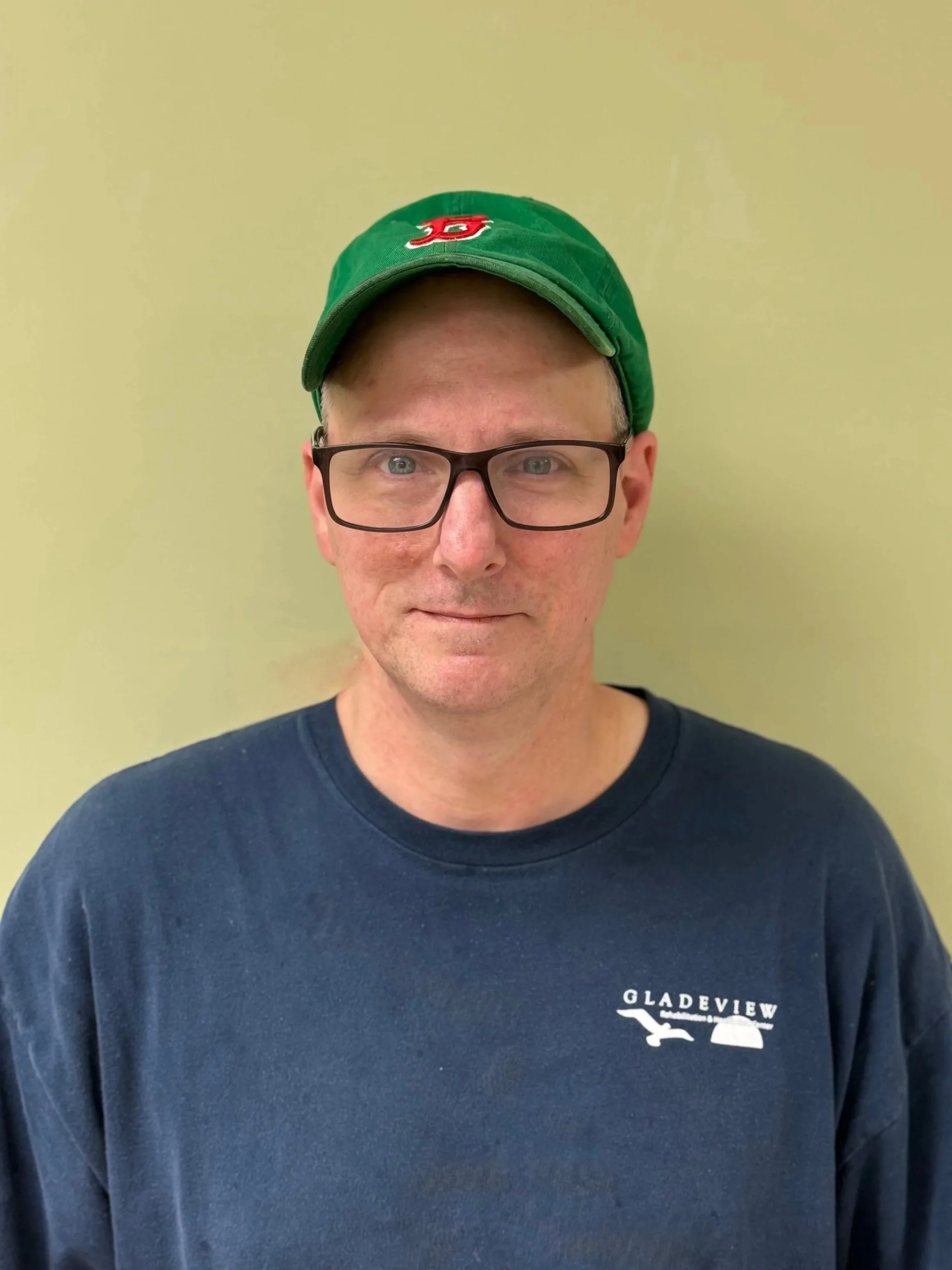 A man with glasses wearing a green baseball cap and a navy blue shirt standing against a yellowish wall.