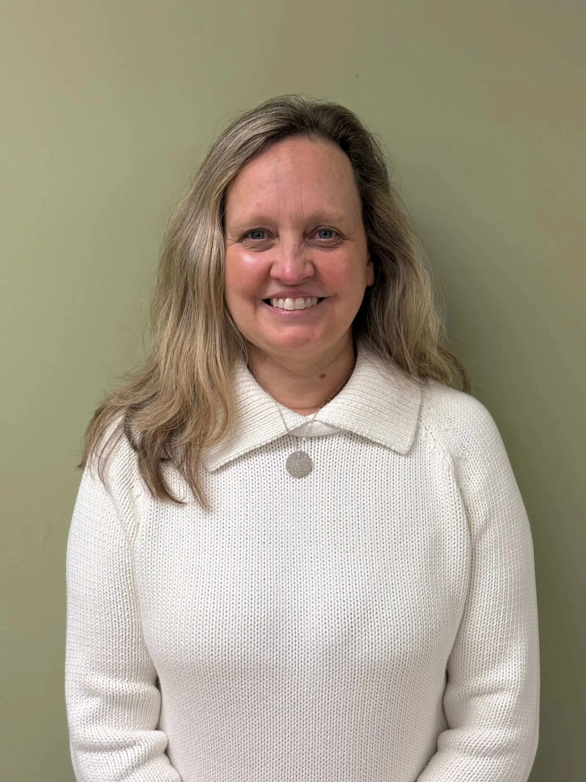 A smiling woman with shoulder-length blonde hair, wearing a cream knit sweater and necklace, standing in front of a plain beige wall.
