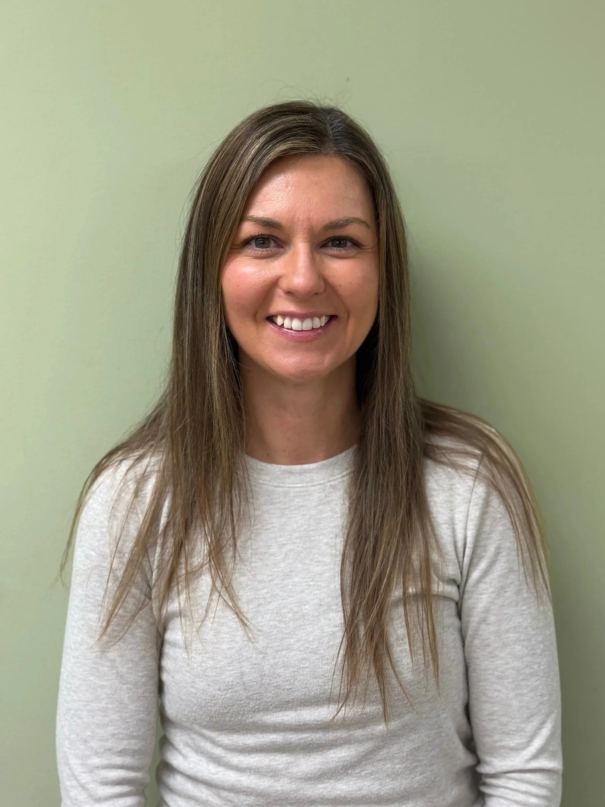 A smiling woman with long brown hair, wearing a white long-sleeve shirt, standing against a light green wall.