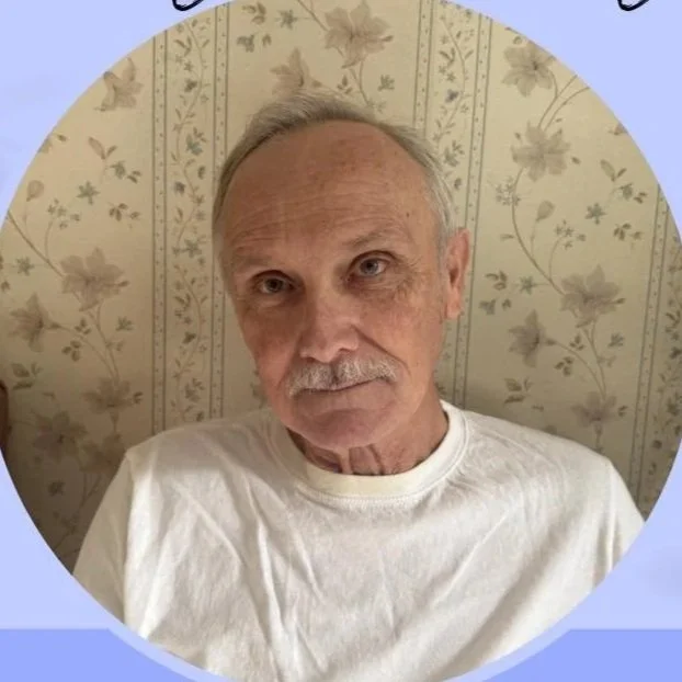 A close-up photo of an elderly man with a mustache and light-colored hair, wearing a white shirt, sitting against a wallpapered background with floral patterns.