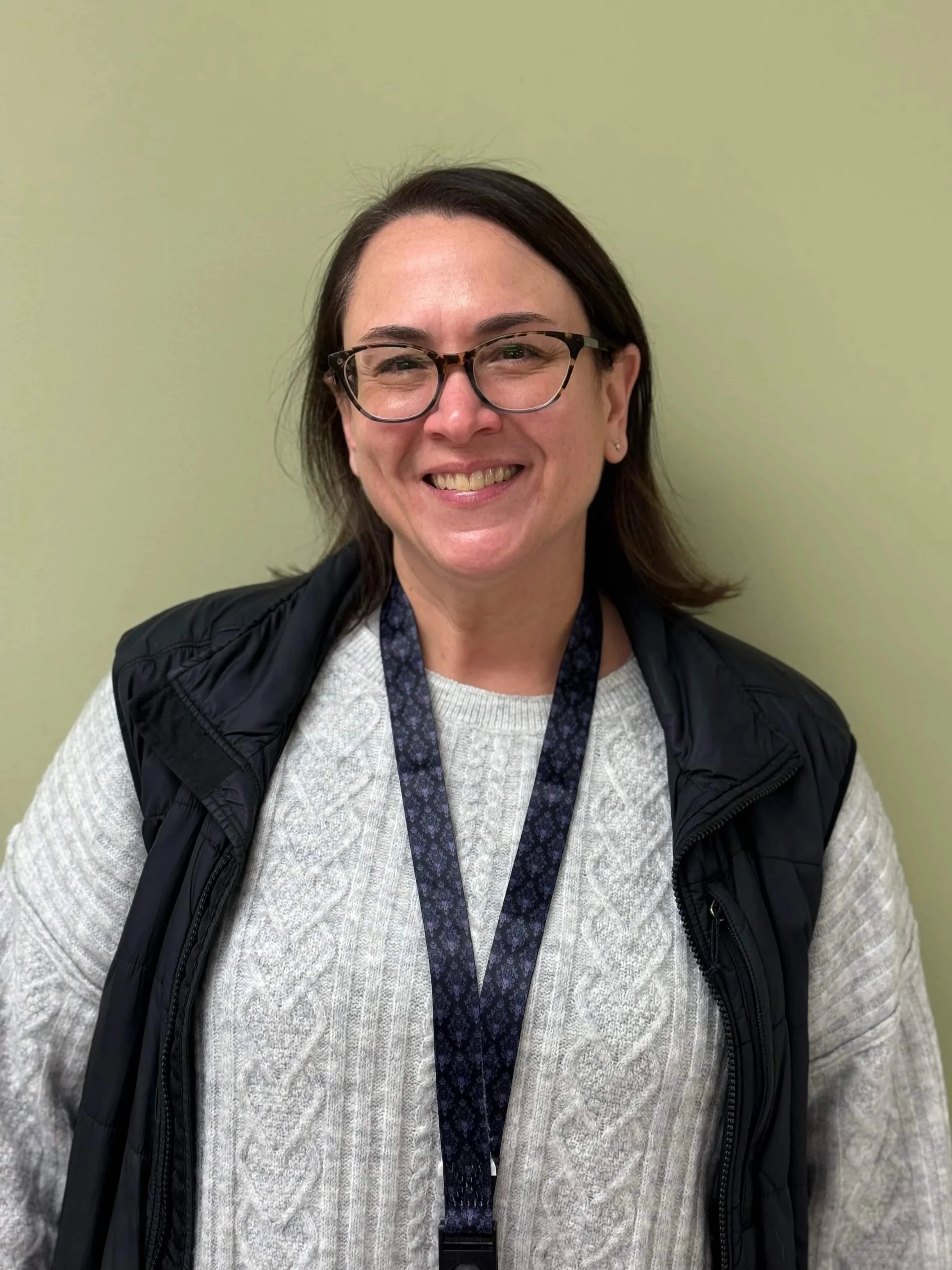 A smiling woman with glasses, dark hair, wearing a light-colored sweater, a black vest, and a lanyard, standing against a plain light green wall.