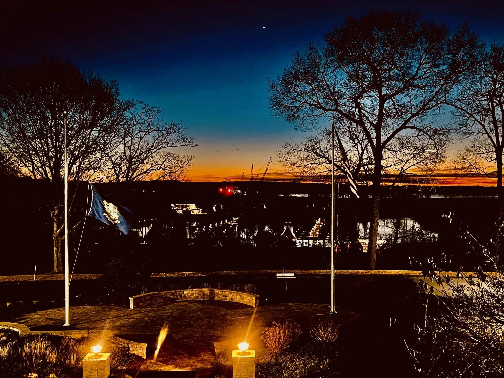 Sunset over a view of trees, water, and a bridge, with flags and lights illuminating a park area in the foreground.