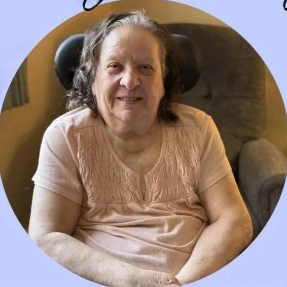 An elderly woman sitting in a chair inside a room, smiling at the camera, with a light pink short-sleeve shirt and dark, curly hair.