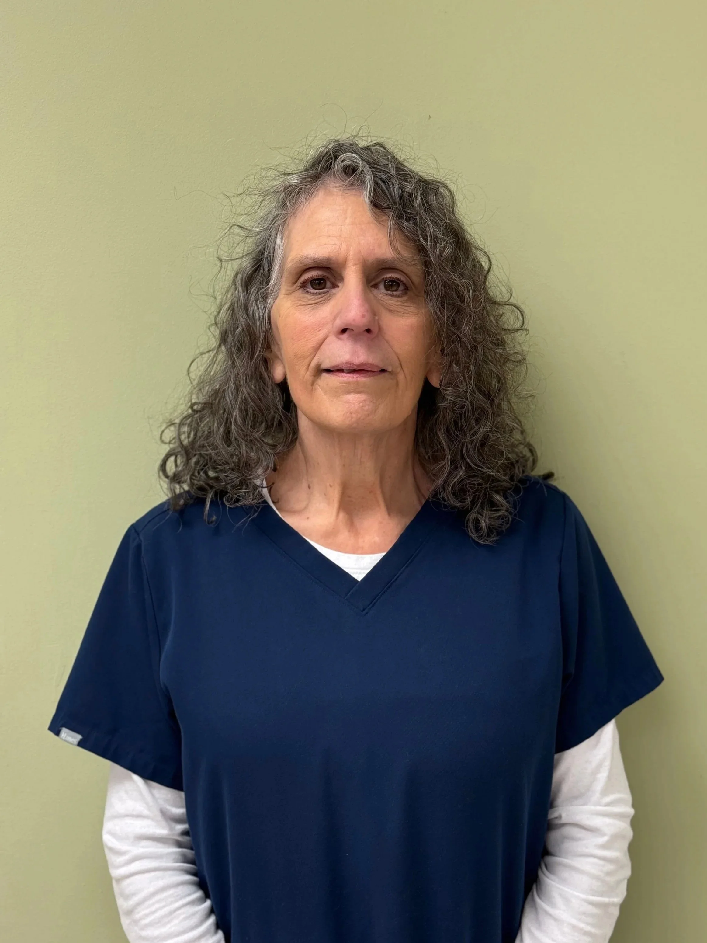 A woman with curly gray hair wearing dark blue scrubs standing against a light green wall.