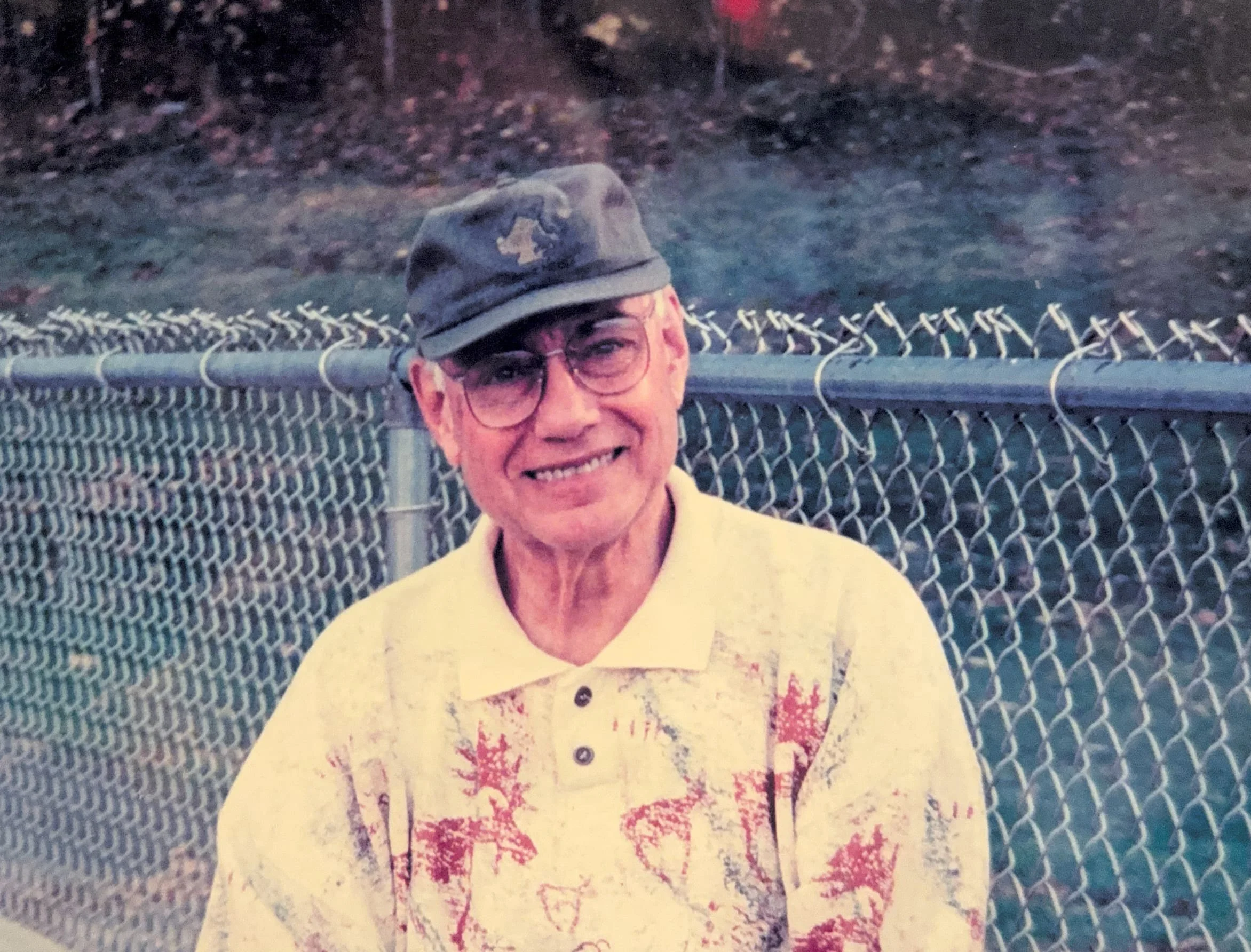 An elderly man wearing glasses, a dark cap, and a beige shirt with red and orange leaf patterns, smiling and standing in front of a chain-link fence with a wooded background.