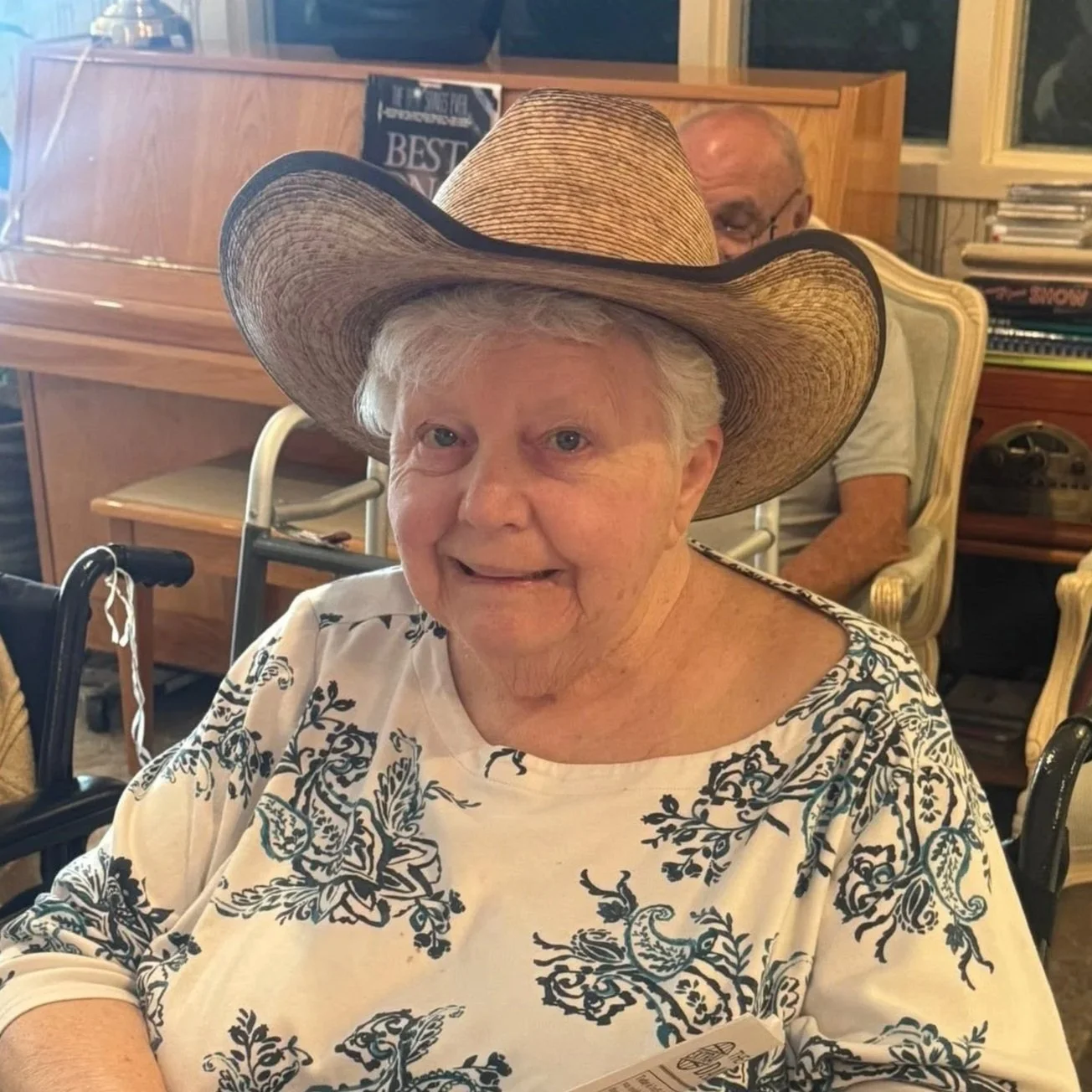 Older woman wearing a large straw cowboy hat and a white shirt with black floral design, sitting indoors at a dining table, smiling at the camera.