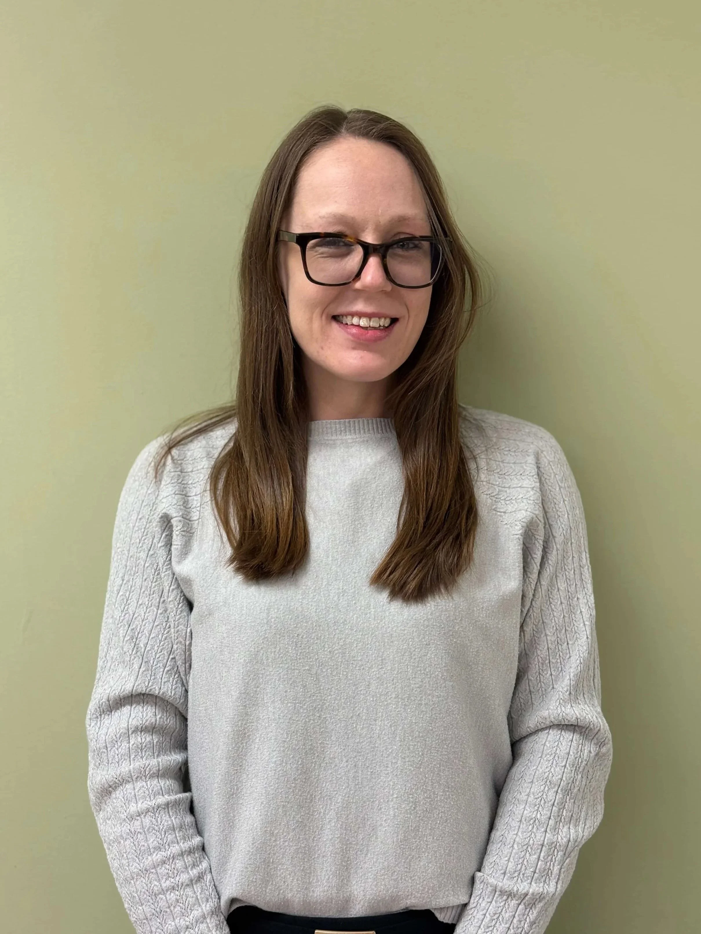 A woman with long brown hair, glasses, and a light gray sweater standing against a light green wall, smiling.