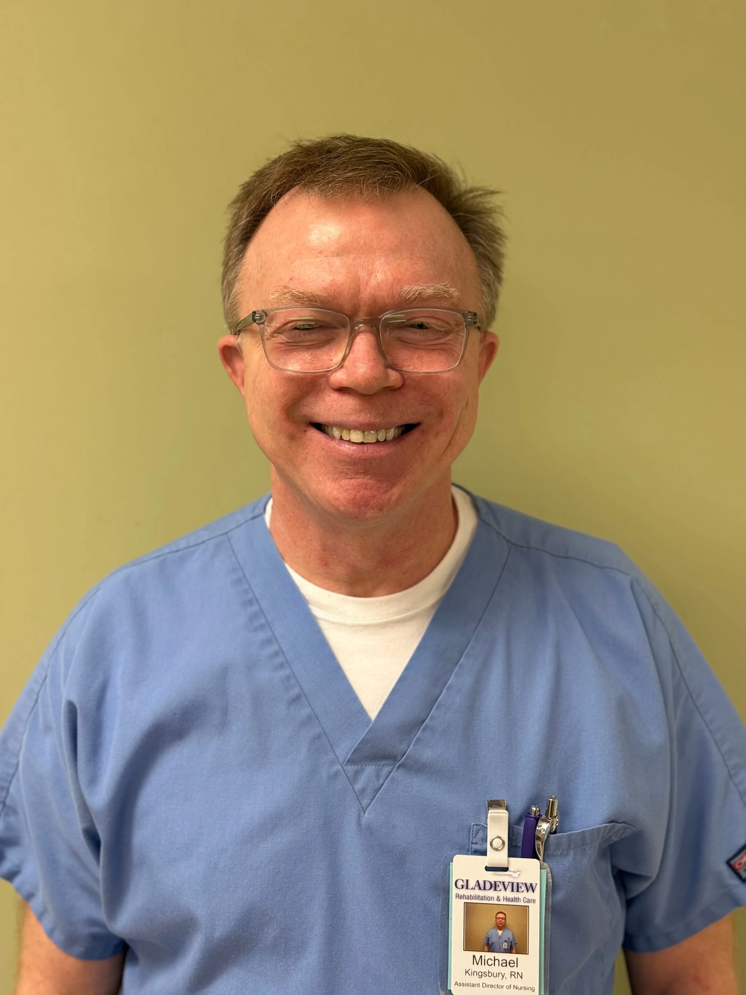 A smiling man in blue medical scrubs with glasses and a name badge, standing against a yellow-green wall.