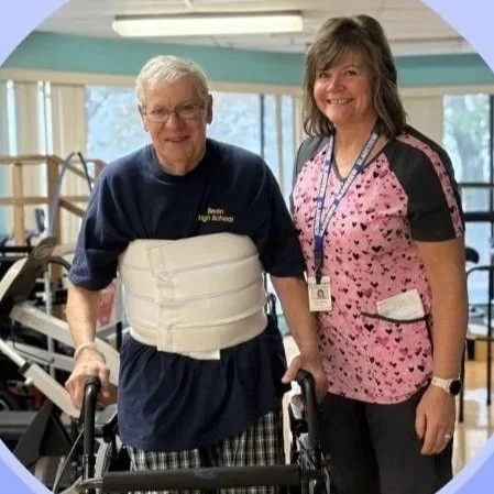 An elderly man with a walker and a supportive brace around her waist standing next to a smiling healthcare worker in pink scrubs inside a rehabilitation facility.