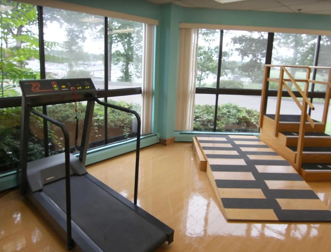 Exercise room with treadmill and wooden stairway, large window views of trees and outdoors, and a patterned black and beige rug.