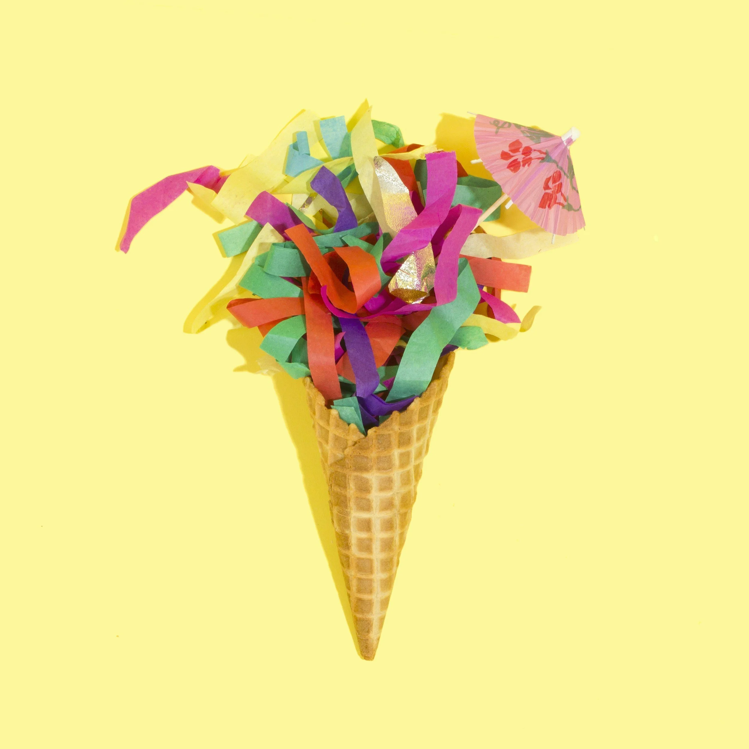An ice cream cone filled with colorful paper streamers and a small paper umbrella on a yellow background.