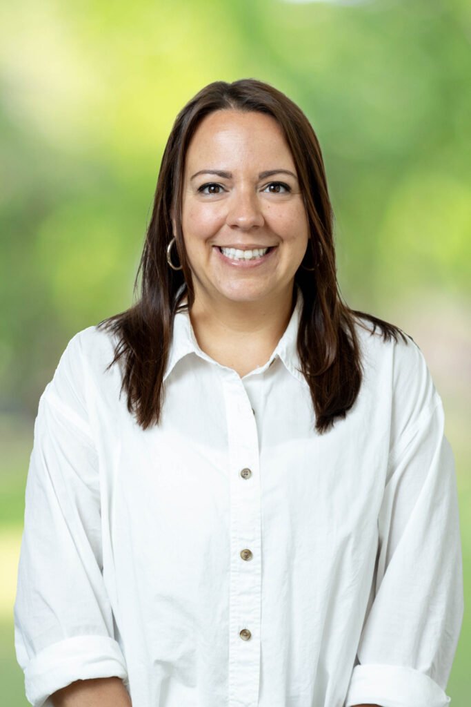 Woman with shoulder-length brown hair, wearing a white button-up shirt, smiling, against a blurred green outdoor background.