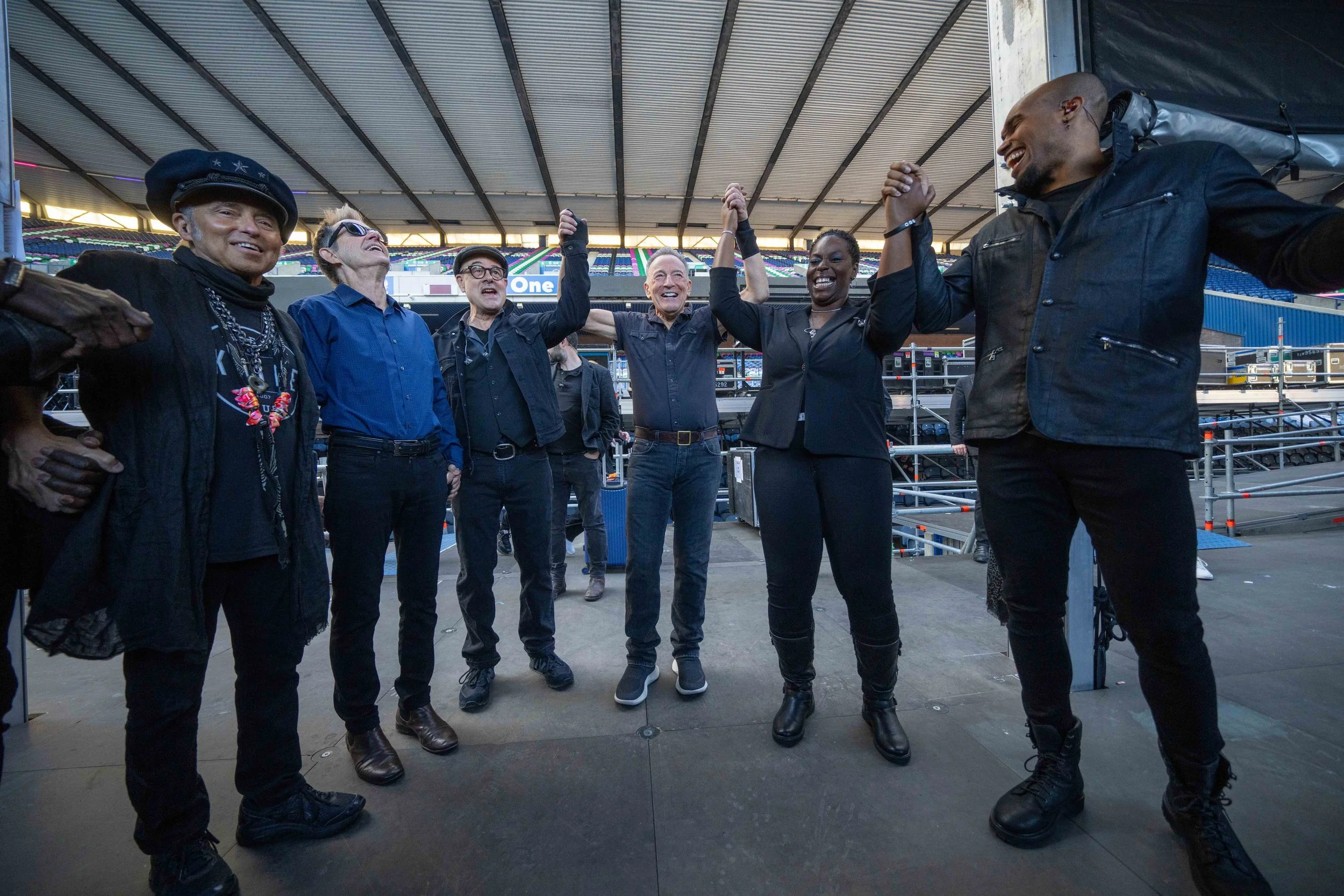 A group of seven diverse people standing on a stage inside a stadium, holding hands in the air and smiling, celebrating happily.