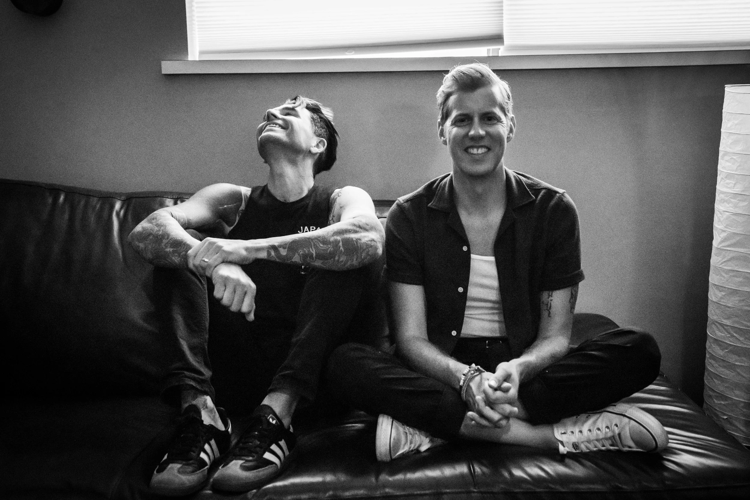 Two young men sitting on a black leather couch, smiling. One man on the right is facing forward with crossed legs and a big smile. The other man on the left is turned to the side, looking up and laughing, with tattoos visible on his arms.