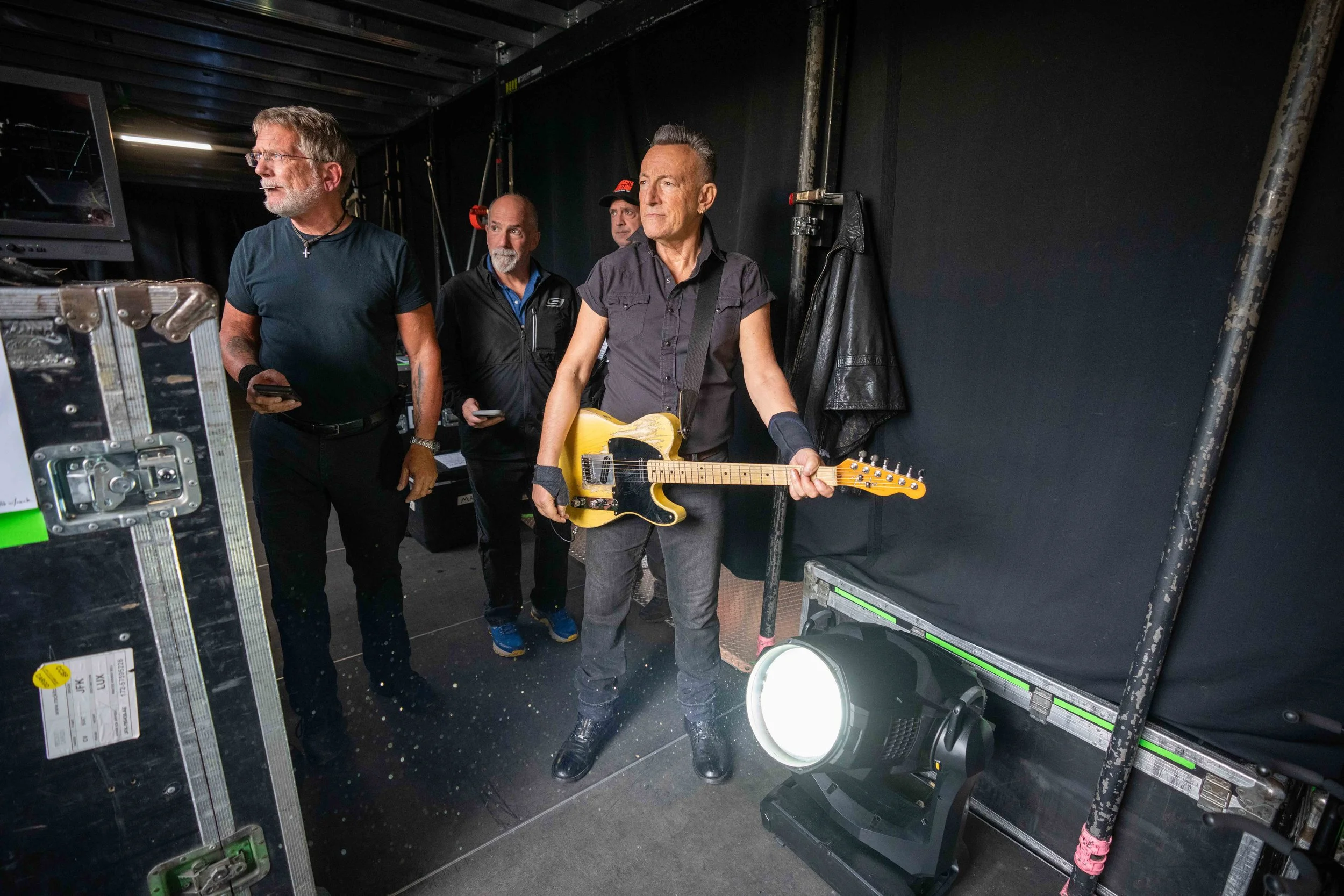 Four men backstage at a concert, one playing an electric guitar and three watching or preparing, with stage equipment and black curtains in the background.