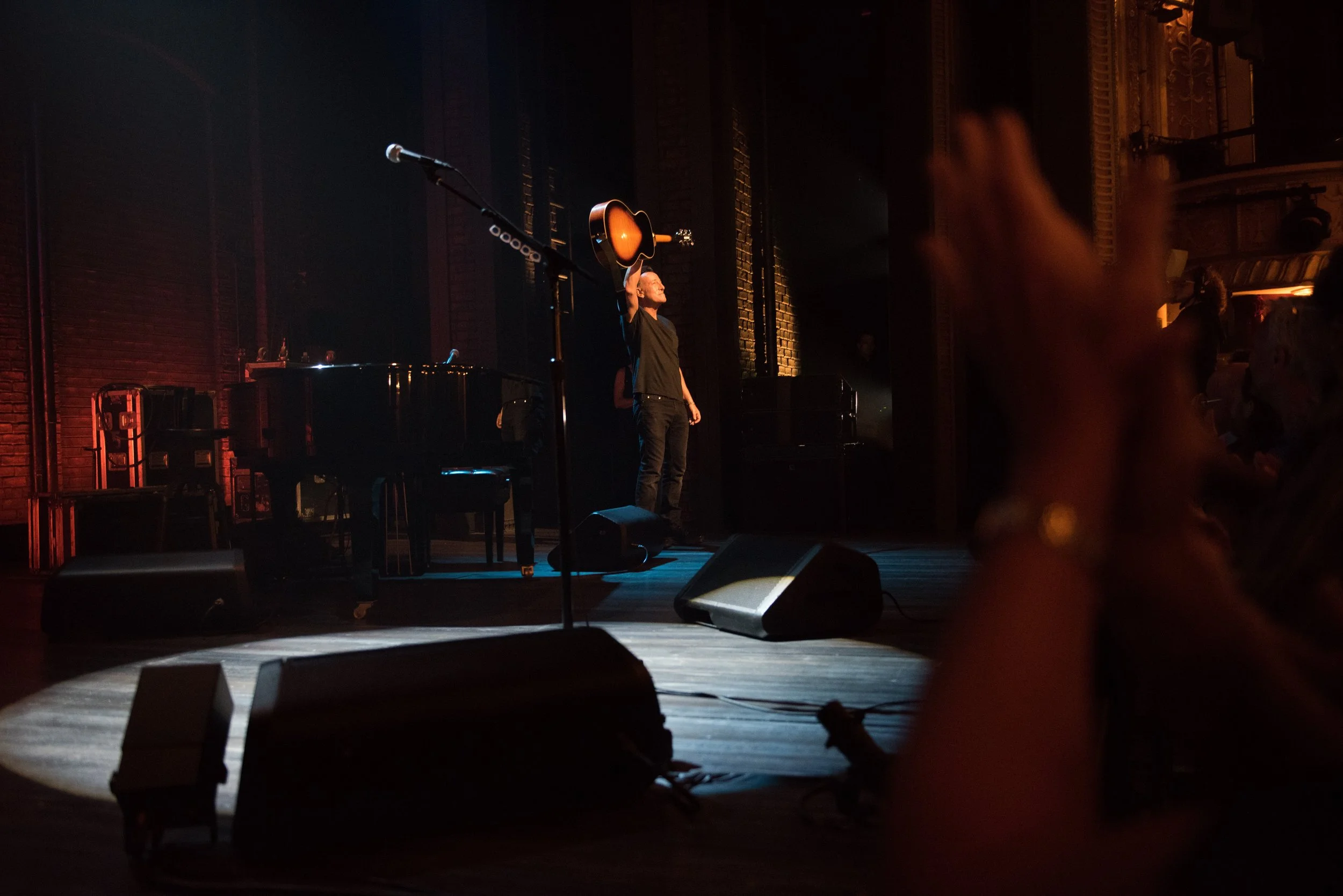 A performer standing on stage with a guitar, taking a bow, in a dimly lit venue with a grand piano and stage monitors. Audience members are visible in the foreground, clapping.