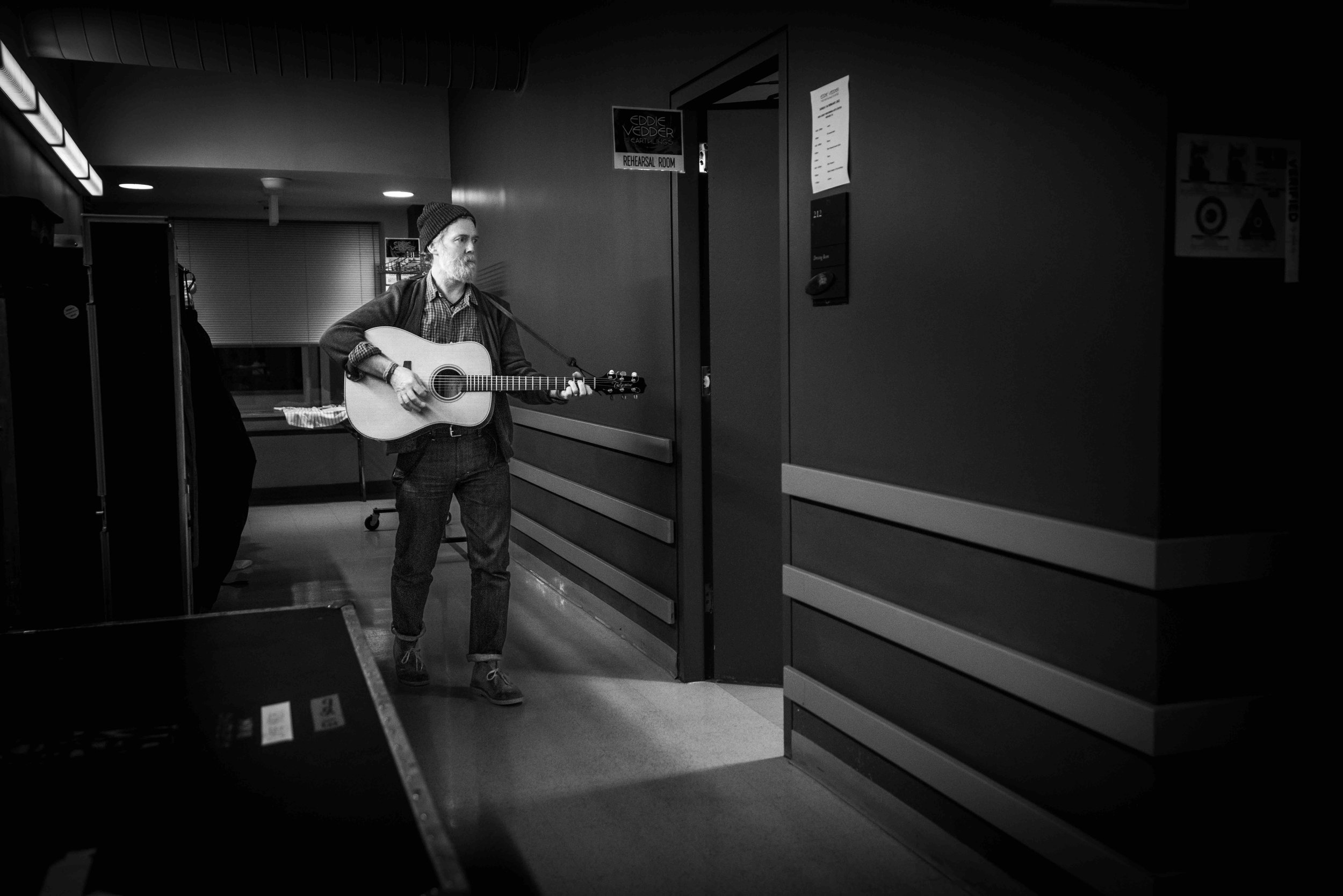 A man wearing a beanie hat and casual clothing, playing an acoustic guitar in a rehearsal room.