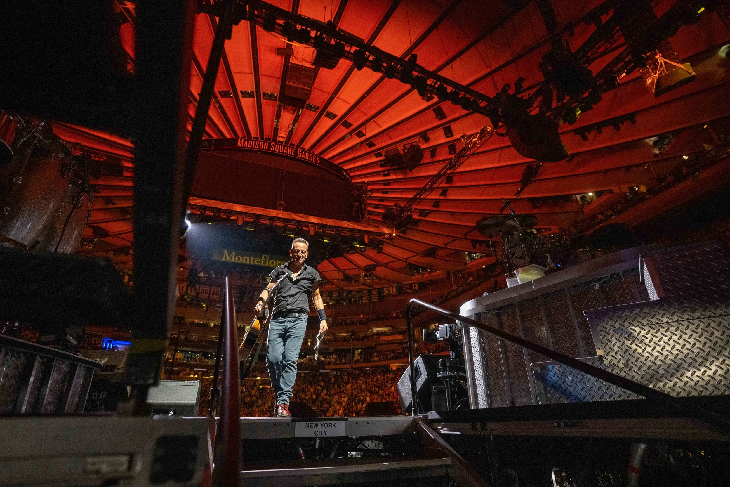 A man walking on a stage with lighting and equipment in front of a large audience in Madison Square Garden, New York City.