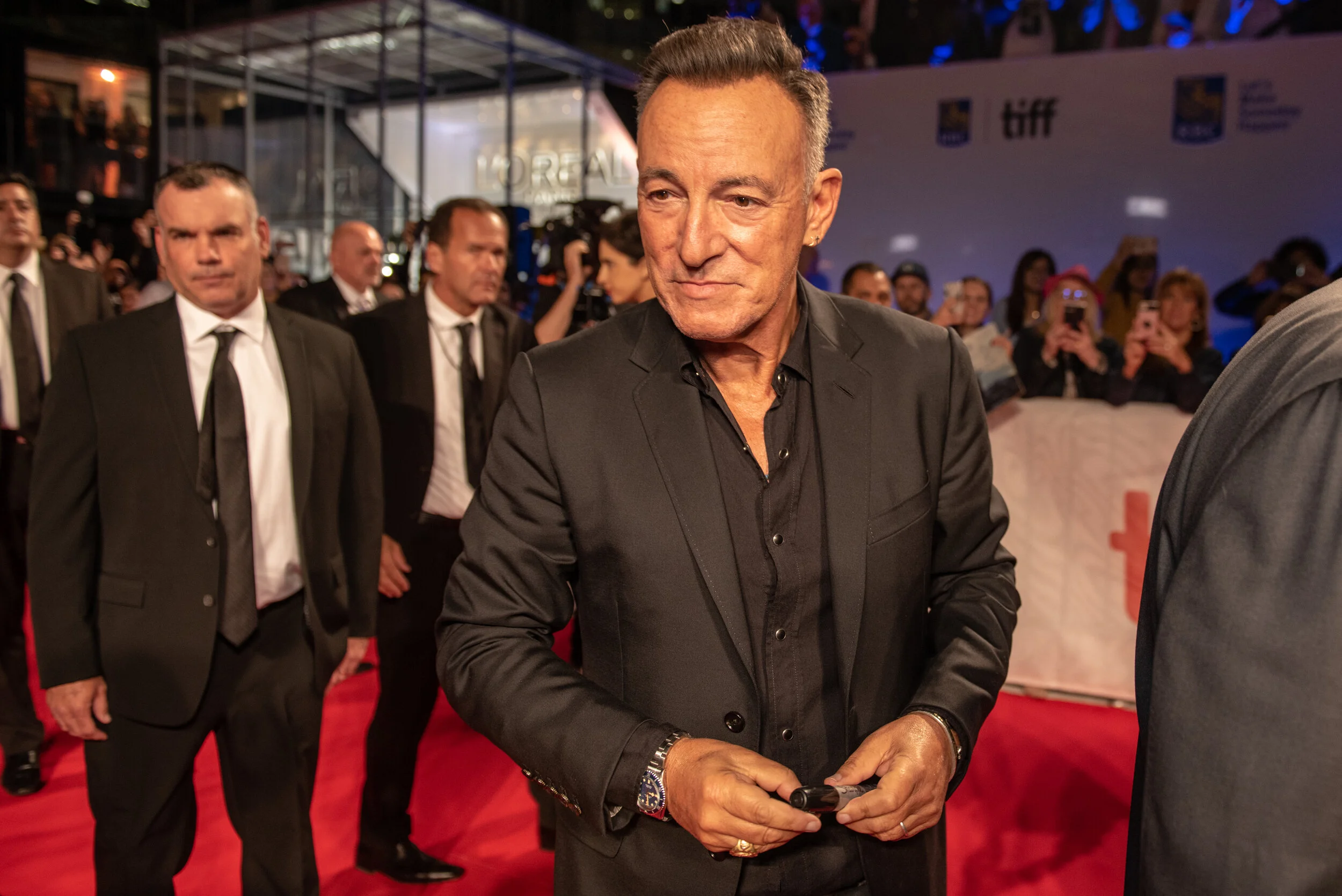 Sean Penn in a black suit at a red carpet event, surrounded by security and fans taking photos.