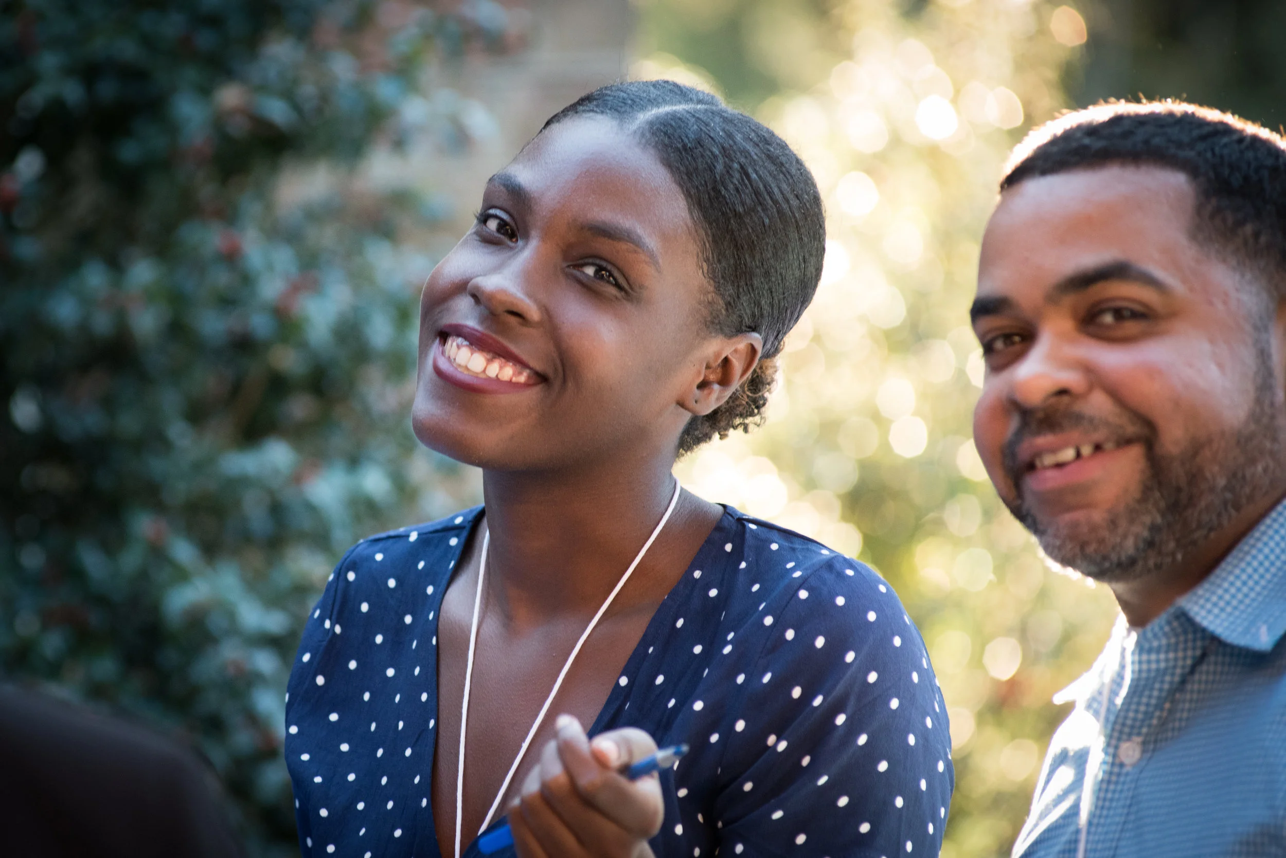 Smiling woman with short hair wearing a polka dot dress, holding a pen, standing outdoors with sunlight and greenery in background, next to a smiling man with beard and short hair wearing a checkered shirt.