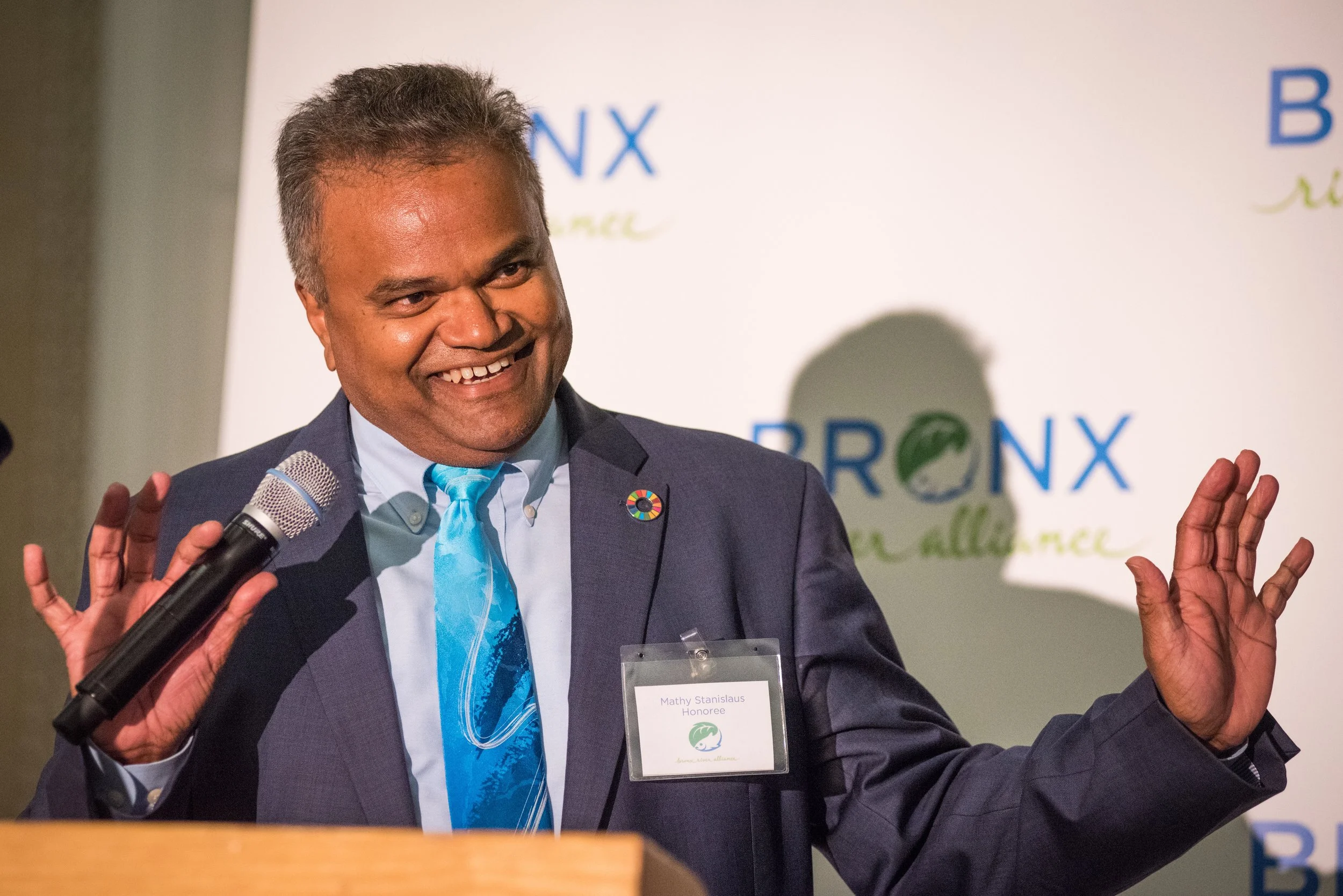 A man with dark hair, wearing a suit and tie, smiling and speaking into a microphone at a conference. He has a name tag that reads 'Mathy Stanislaus Honoree' and a circular pin on his lapel. Behind him is a banner with the words 'BRNX' and 'resilienc