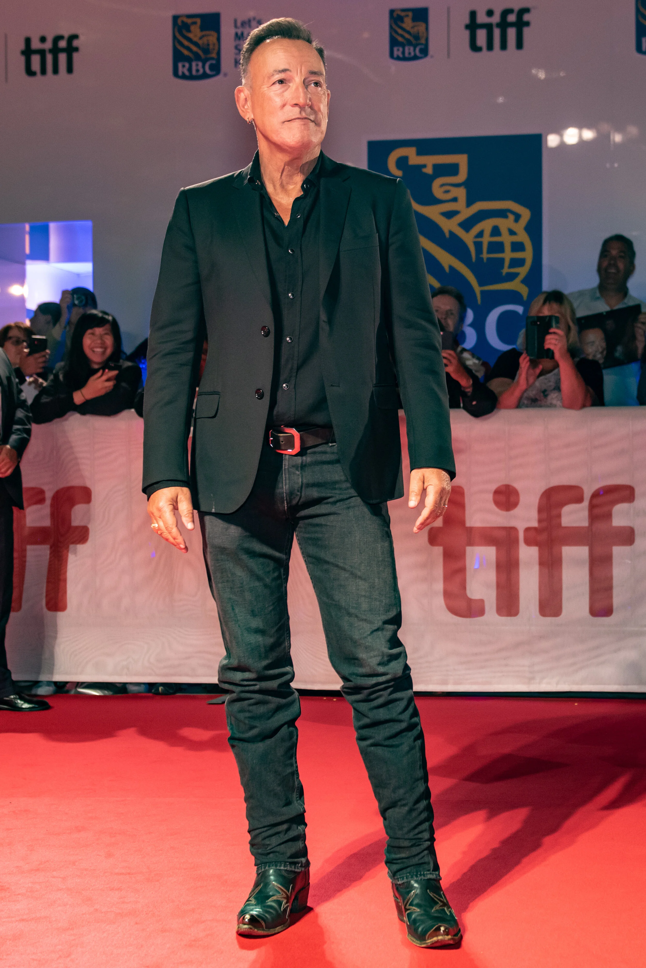 A man wearing a black blazer, black shirt, and black jeans standing on a red carpet at a TIFF event, with fans in the background taking photos and a TIFF banner visible.