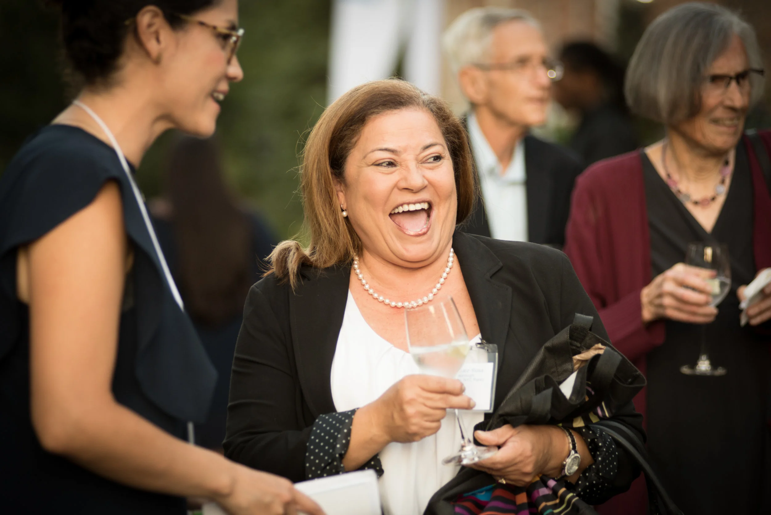 A group of middle-aged adults at an outdoor social event, engaging in conversation, with one woman laughing and holding a glass of wine while wearing a pearl necklace.