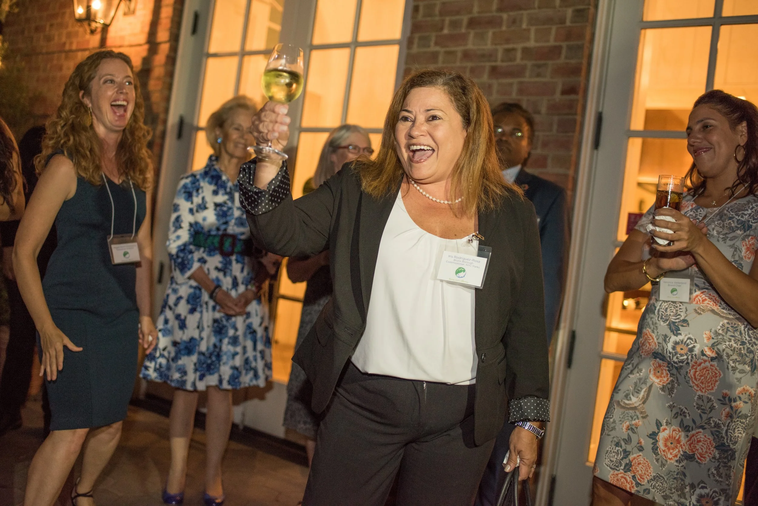 Group of women at a social event, with one woman in the center raising a glass of white wine, smiling and wearing a black blazer and white blouse, surrounded by others holding drinks and smiling.