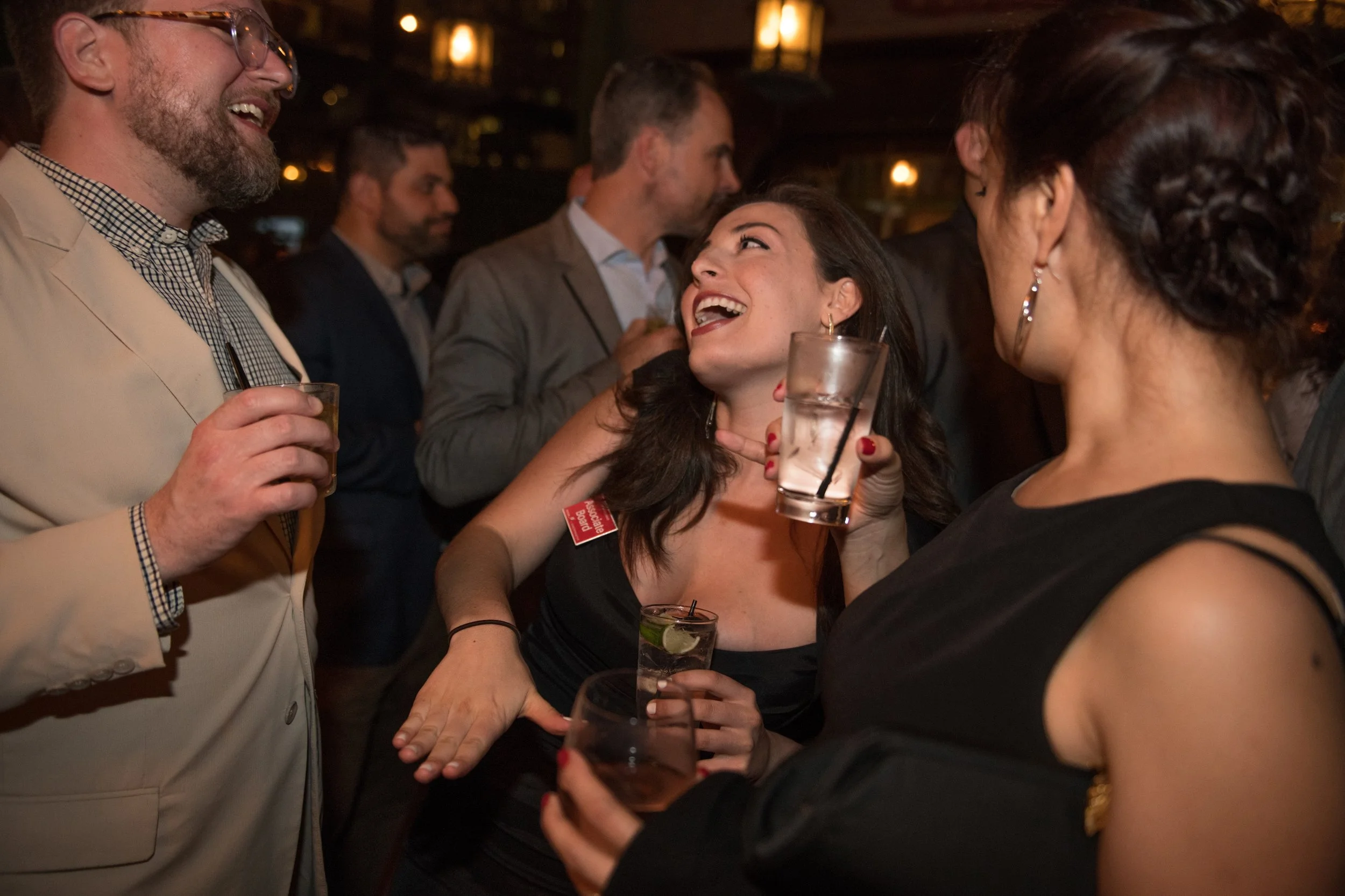 Group of people socializing, smiling, and holding drinks at a lively party or bar, with warm lighting and a dark background.