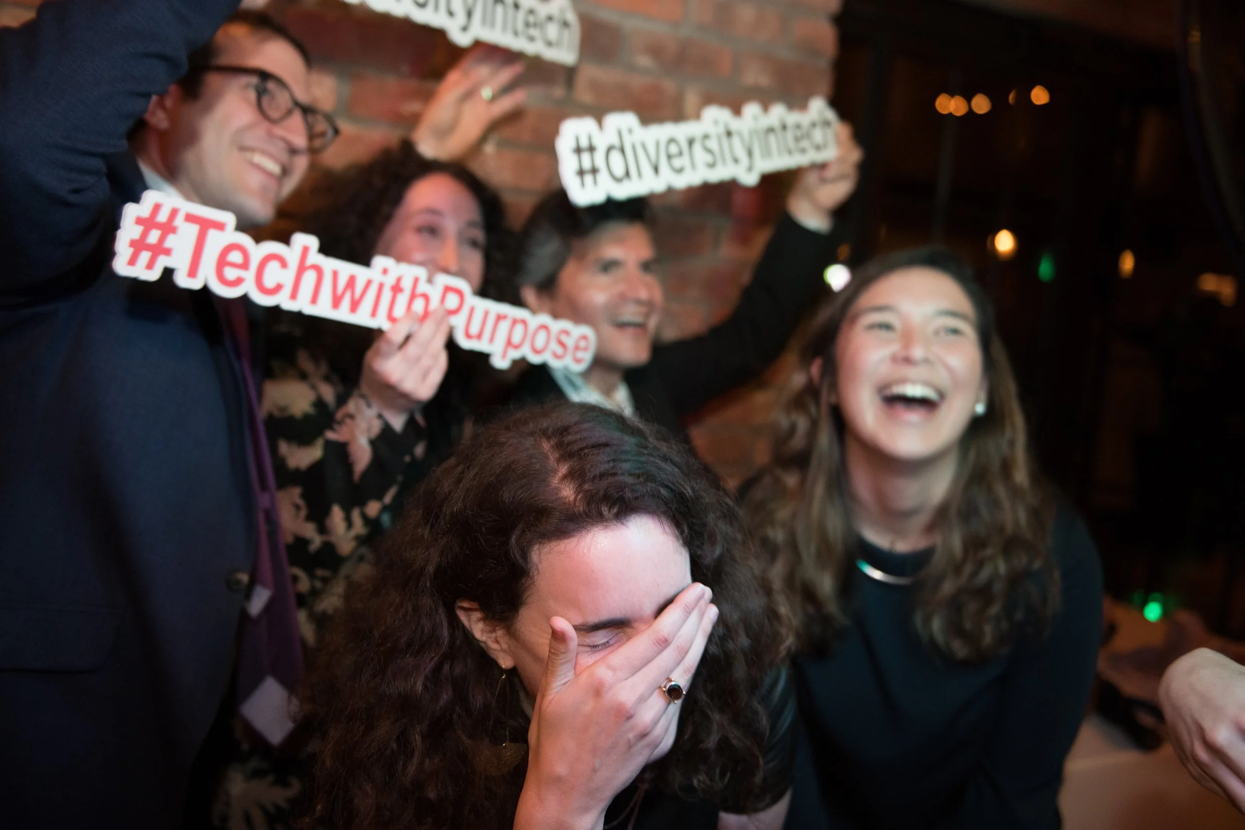 Group of five people celebrating and laughing together indoors, holding signs that read '#DiversityInclusion', '#TechwithPurpose', and '#DiversityInclusion', with a brick wall in the background.